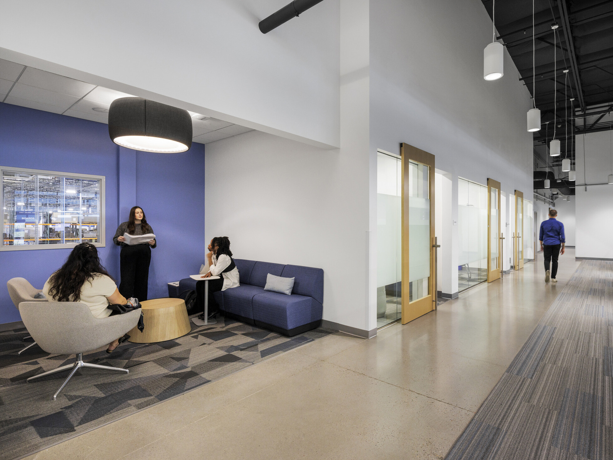 Sub-Zero Group work space corridor; small gathering spot at left with three employees in conversation; warehouse-facing window connecting office and production floor