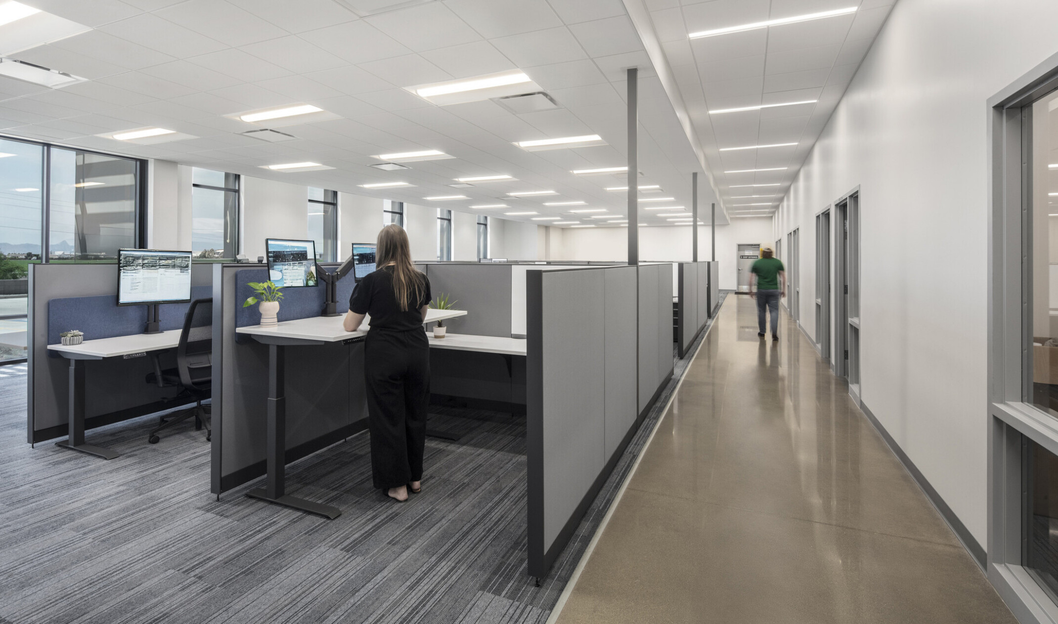 Wildeck workspace; window-lined workstations with seated-height privacy walls and standing desks; offices and conference rooms positioned along the interior, separated by a corridor; floor-to-ceiling windows bringing in natural light