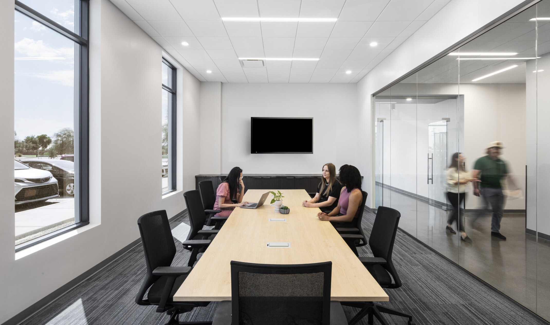 Wildeck conference room; gray-toned carpet with linear pattern; light oak conference table with employees seated in a meeting; glass walls bringing natural light deep into the space