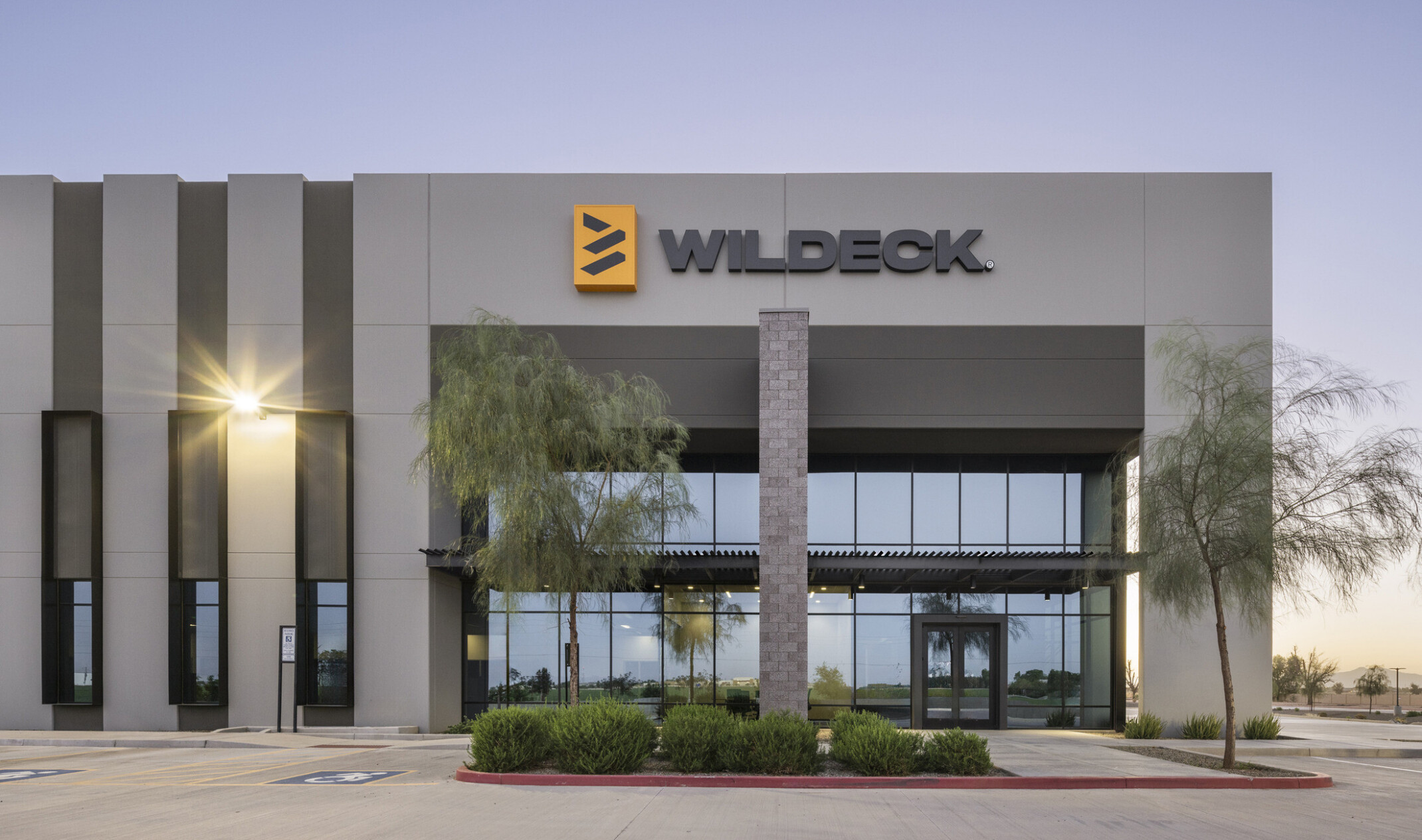 Wildeck building entrance; corner view of glass front facade with black trim, canopy detail, brick accent, and vertical striping detail to the left of the entrance; Wildeck signage displayed above entry