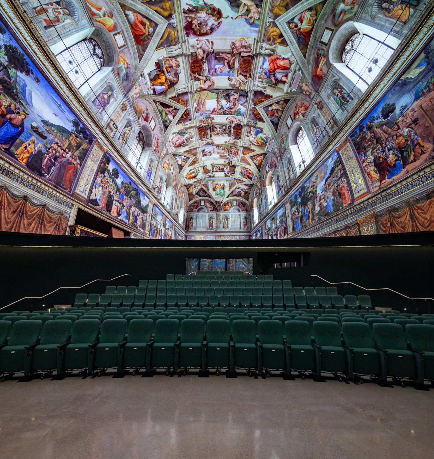 wide-angle view of a hall with rows of green theater-style seats facing a wall, above which is a replica of the Sistine Chapel ceiling. The ceiling and upper walls are covered with vibrant Renaissance frescoes depicting biblical scenes, framed by architectural details. The perspective emphasizes the ornate artwork overhead and the symmetry of the seating below