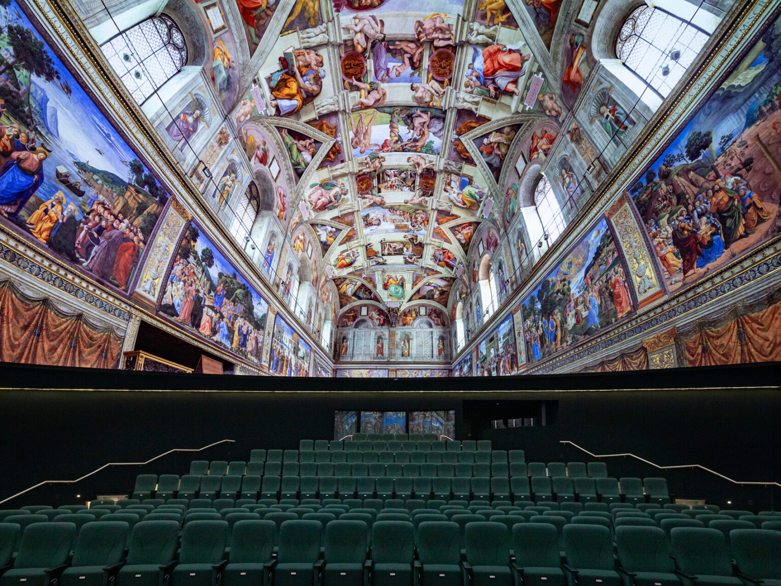 wide-angle view of a hall with rows of green theater-style seats facing a wall, above which is a replica of the Sistine Chapel ceiling. The ceiling and upper walls are covered with vibrant Renaissance frescoes depicting biblical scenes, framed by architectural details. The perspective emphasizes the ornate artwork overhead and the symmetry of the seating below