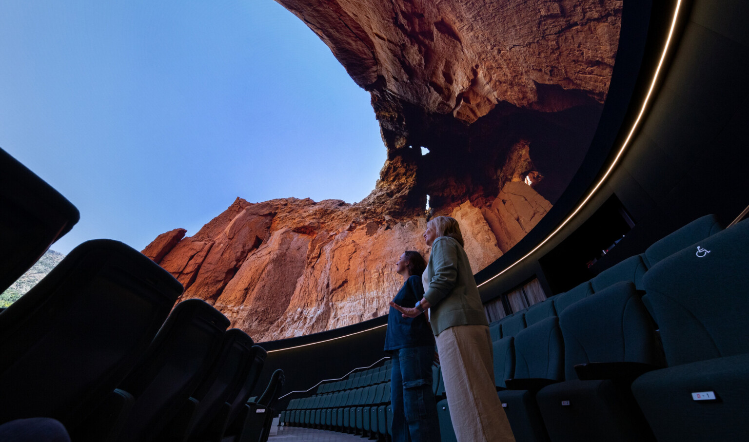 Interior view of a modern theater with curved rows of dark green seats. Two people stand near the front, looking up at a massive curved screen displaying a vivid image of a red rock canyon with a clear blue sky above. The screen wraps around the space, creating an immersive experience