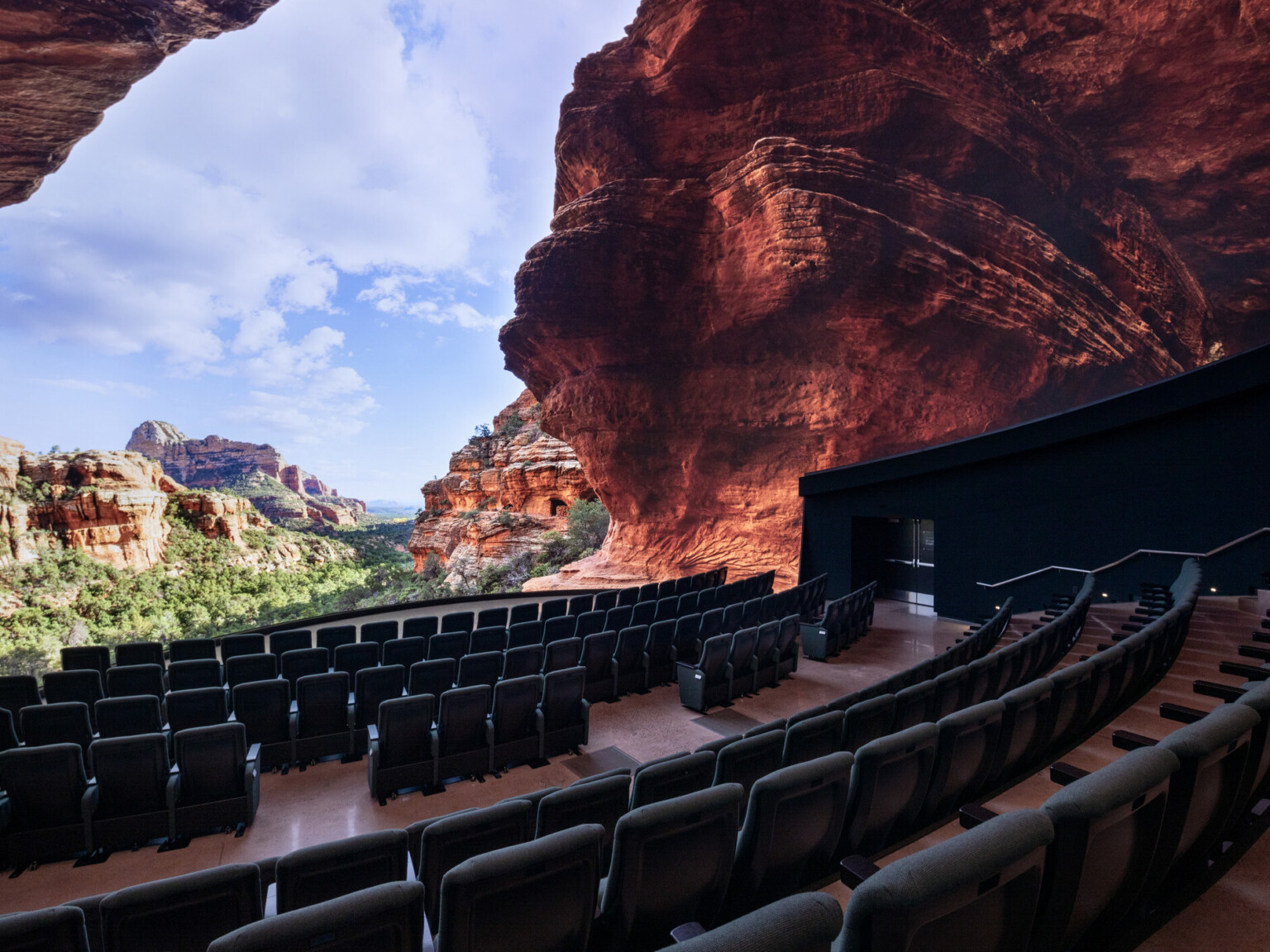 An immersive theater with rows of dark seats facing a massive curved screen that displays a vivid panoramic view of a red rock canyon under a partly cloudy blue sky. The screen wraps around the space, creating the illusion of being inside a natural cave opening overlooking rugged cliffs and greenery