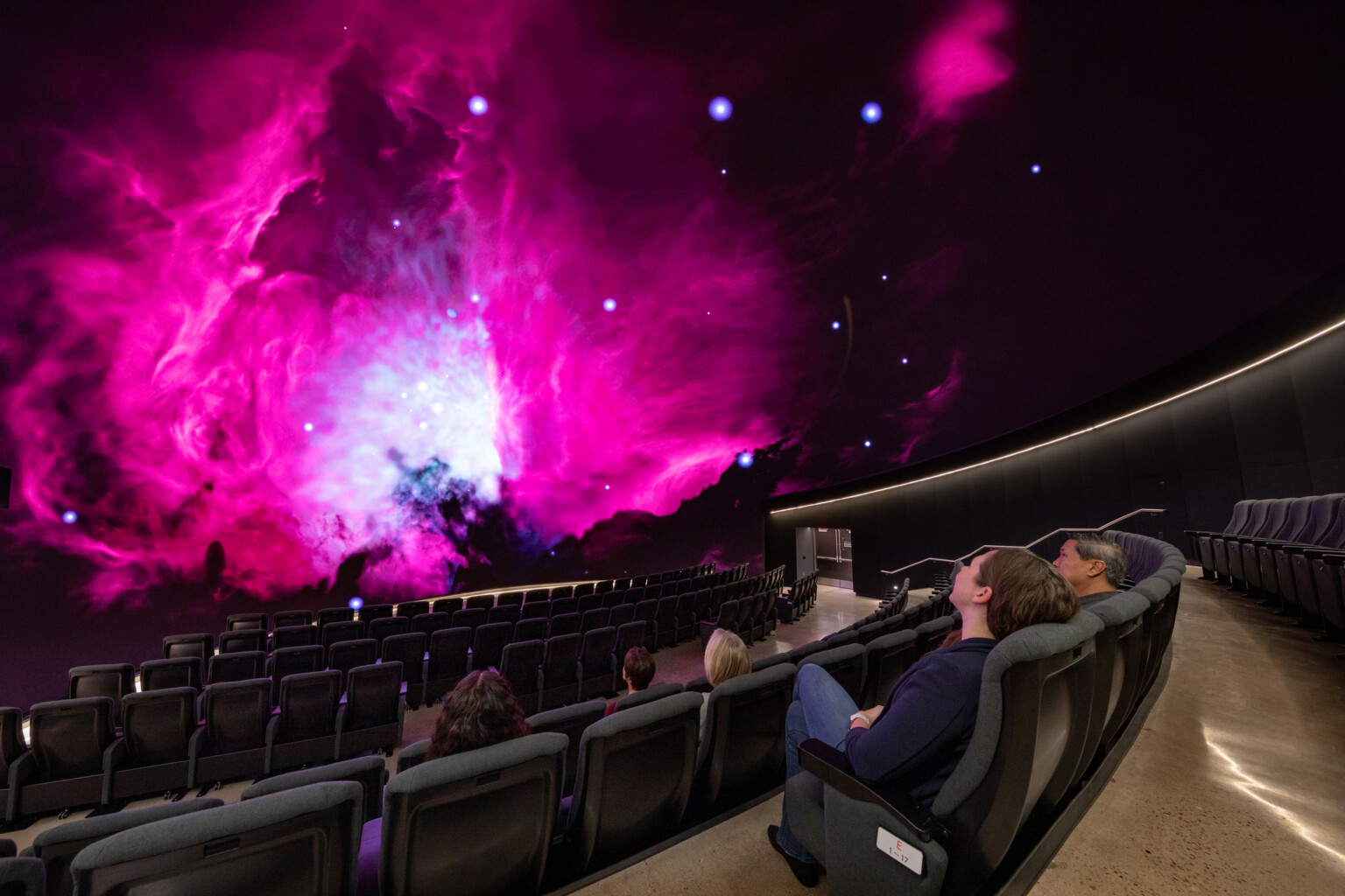 immersive theater with curved rows of seats facing a massive screen that displays a outerspace scape in blue, pink, and purple hues.