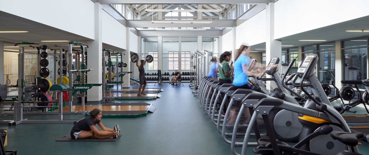 High school gym with high ceilings lined with windows filled with exercise equipment sitting on dark green floors