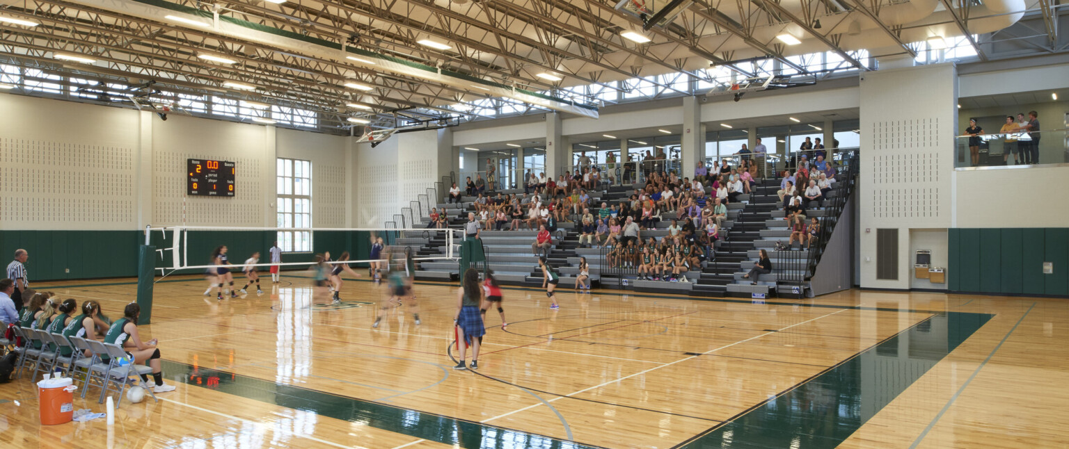 High school basketball gym with grey bleachers, natural wood floors with green accents, exposed ceiling beams with girls playing volleyball