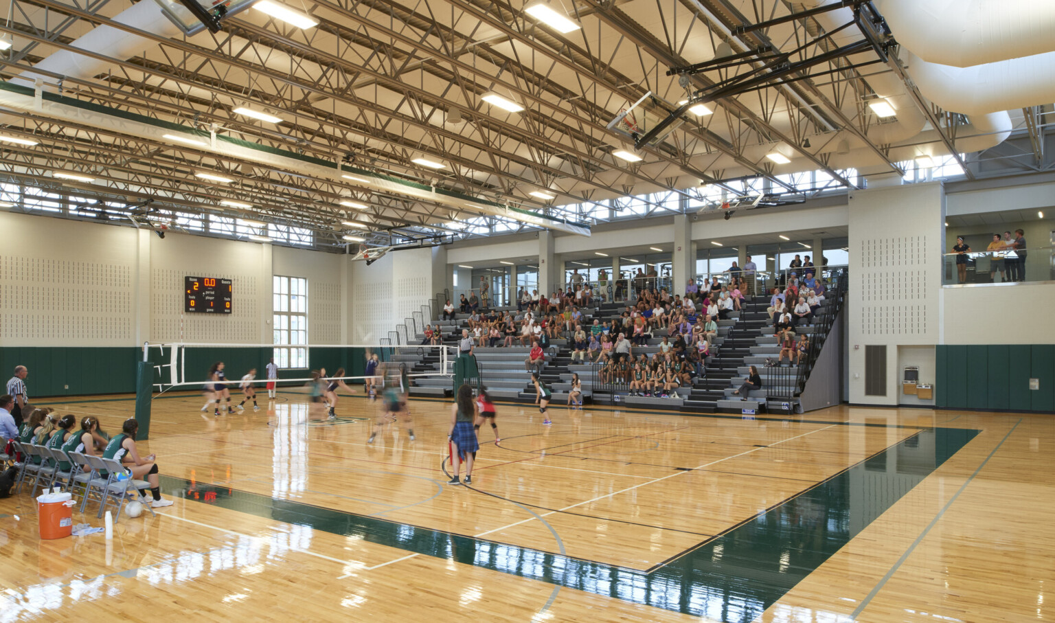 High school basketball gym with grey bleachers, natural wood floors with green accents, exposed ceiling beams with girls playing volleyball
