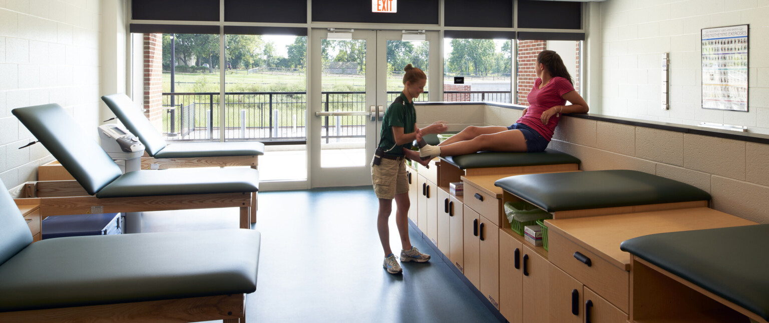 Sports medicine room showing light wood cabinets with green cushions on top. Wall lined with green cushioned exam tables looking out to a wall of windows