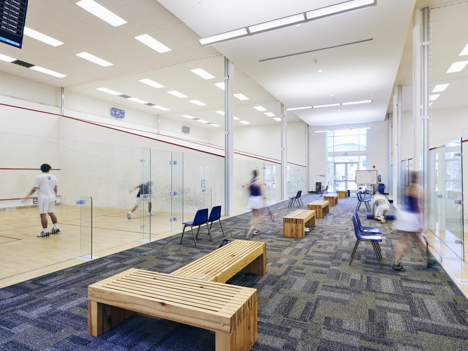 Long hallway filled with wooden benches with glassed in squash courts on both sides