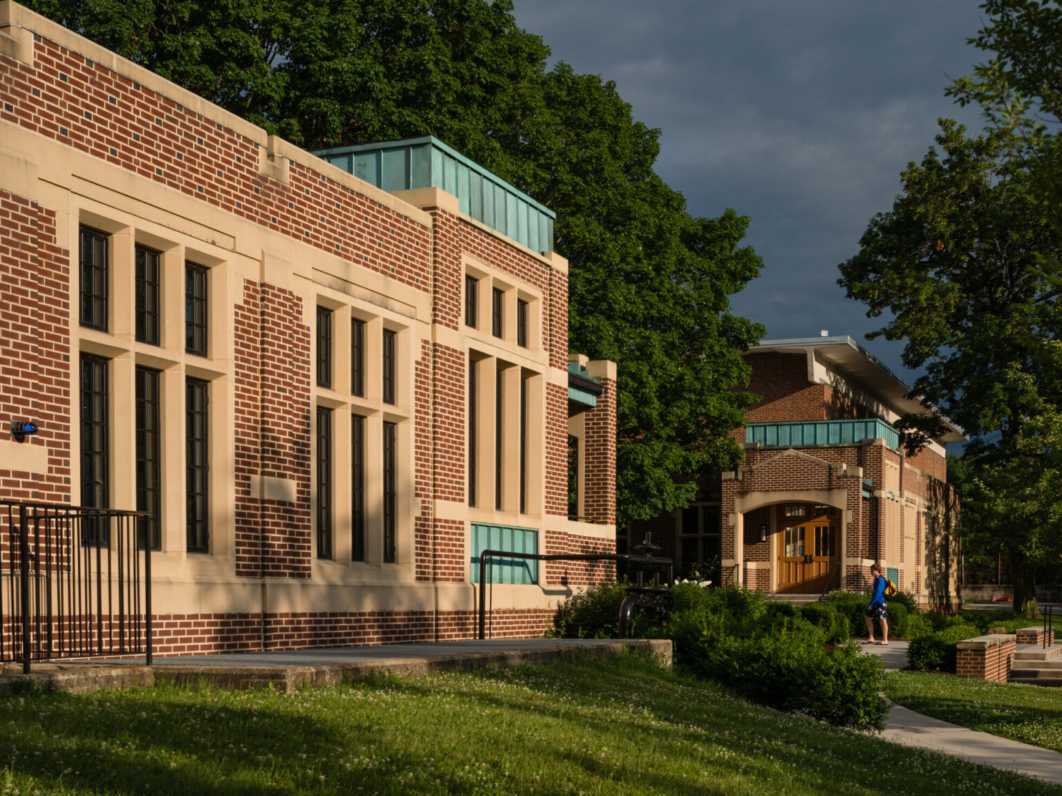Single story brick building with windows outlined in off white with green accents on roofs surrounded by large green trees