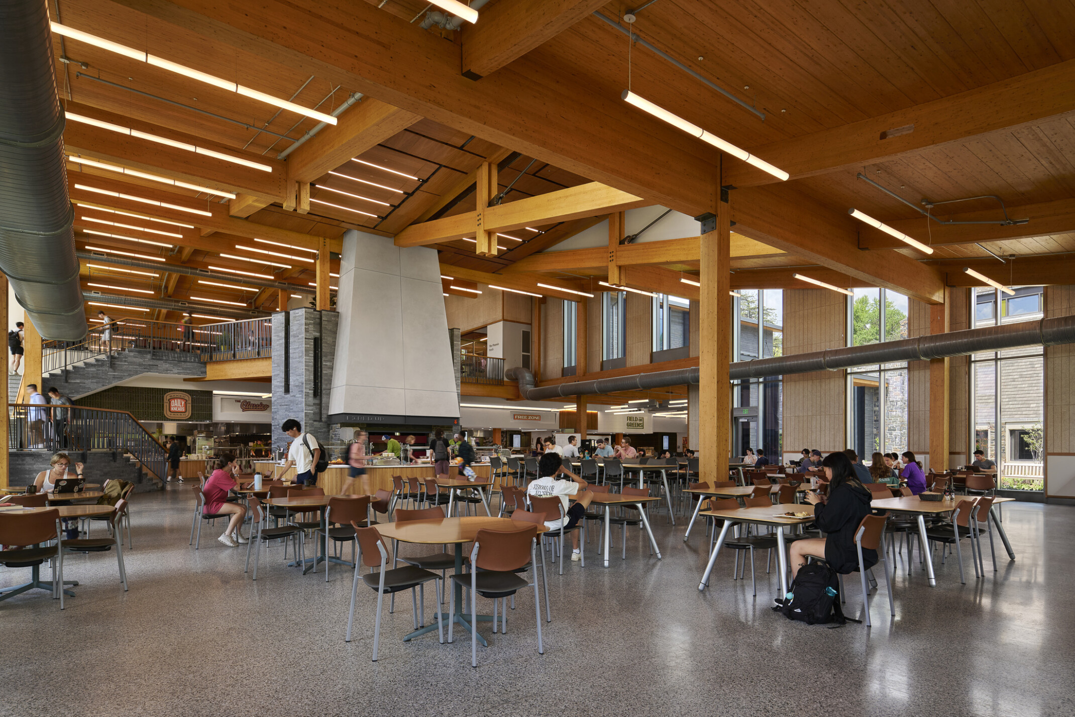 large open cafeteria with multiple storeys with seating for students; Mass timber ceiling makes for a warm and industrial space with exposed stone and venting