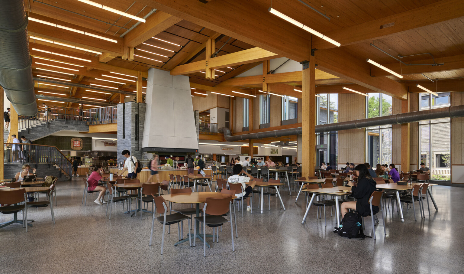 large open cafeteria with multiple storeys with seating for students; Mass timber ceiling makes for a warm and industrial space with exposed stone and venting