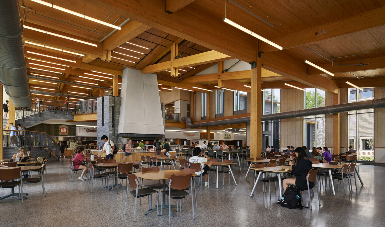 large open cafeteria with multiple storeys with seating for students; Mass timber ceiling makes for a warm and industrial space with exposed stone and venting