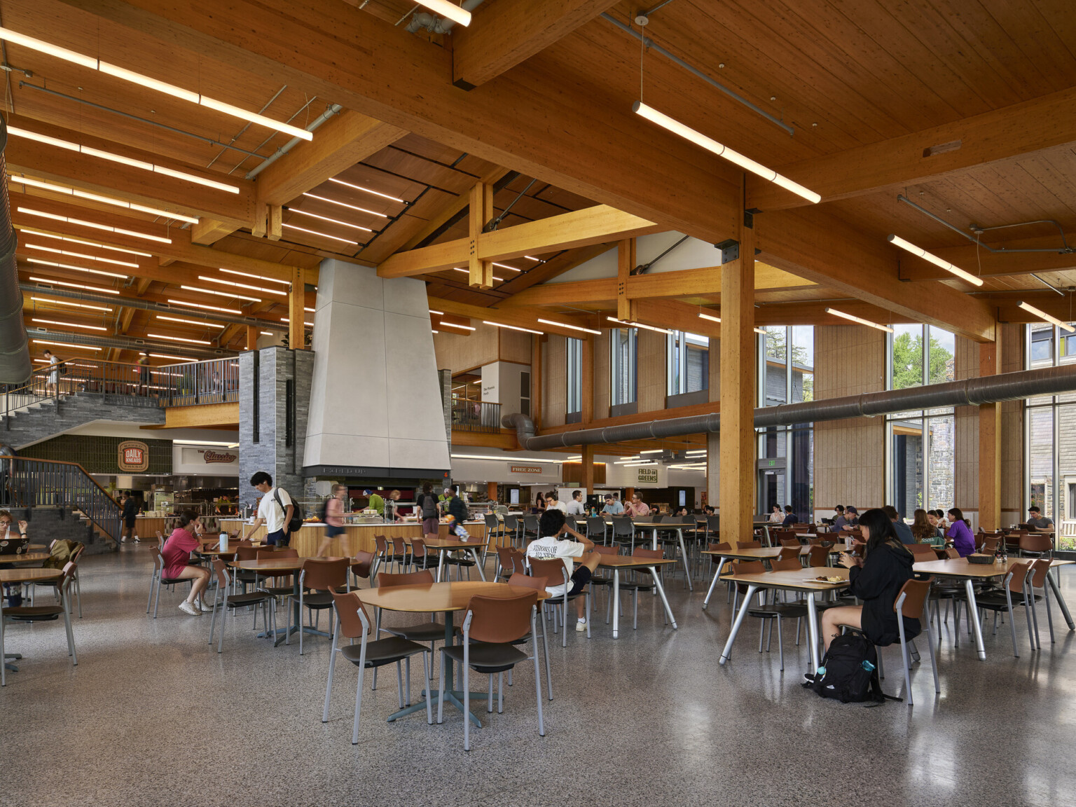 large open cafeteria with multiple storeys with seating for students; Mass timber ceiling makes for a warm and industrial space with exposed stone and venting large open cafeteria with multiple storeys with seating for students; Mass timber ceiling makes for a warm and industrial space with exposed stone and venting