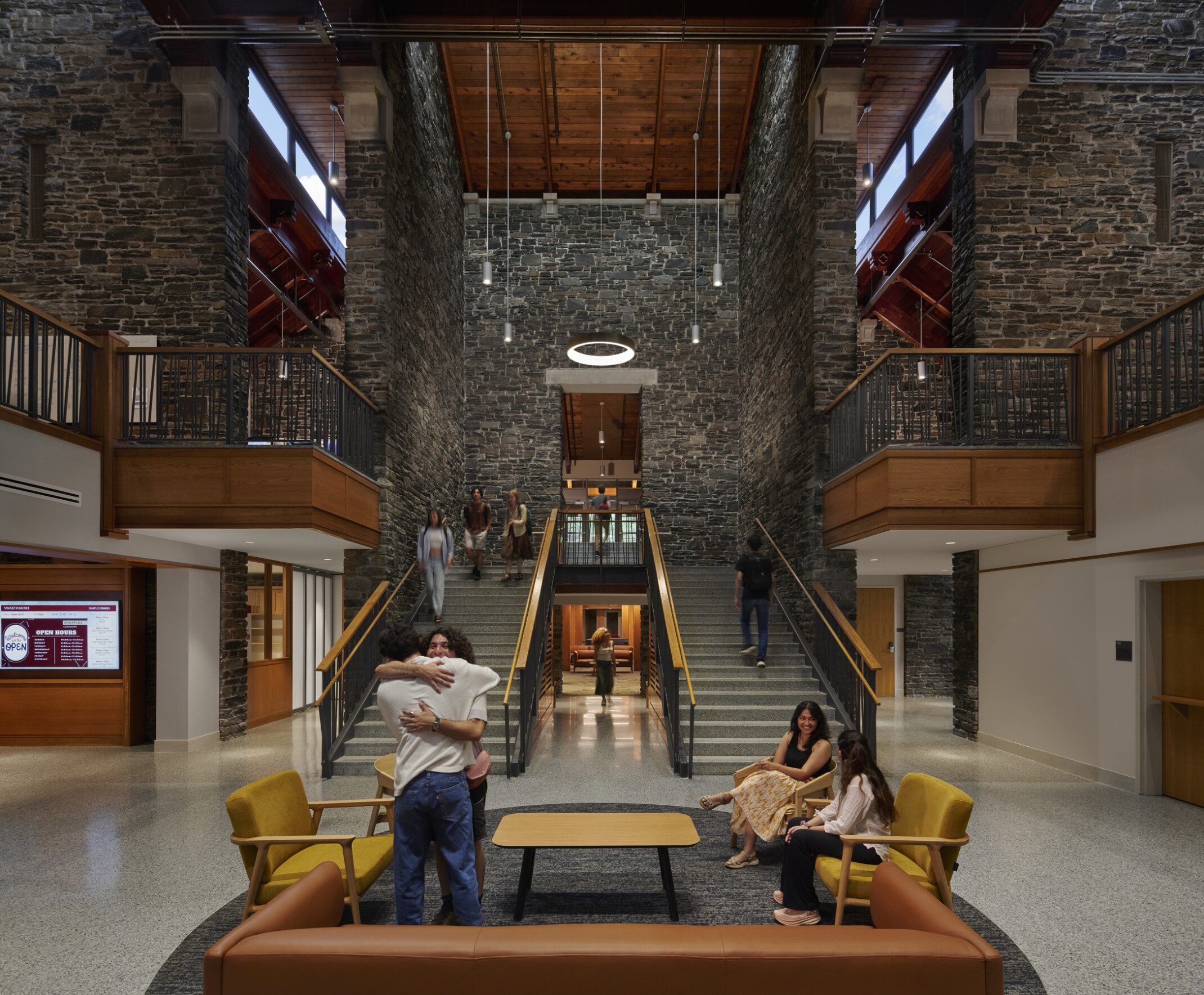 Broad staircase with wooden railings ascending to a second floor, flanked by stone walls, circular light fixture suspended from the ceiling. Seating area with yellow and brown chairs and a rectangular wooden coffee table on a speckled gray floor at the base of the stairs