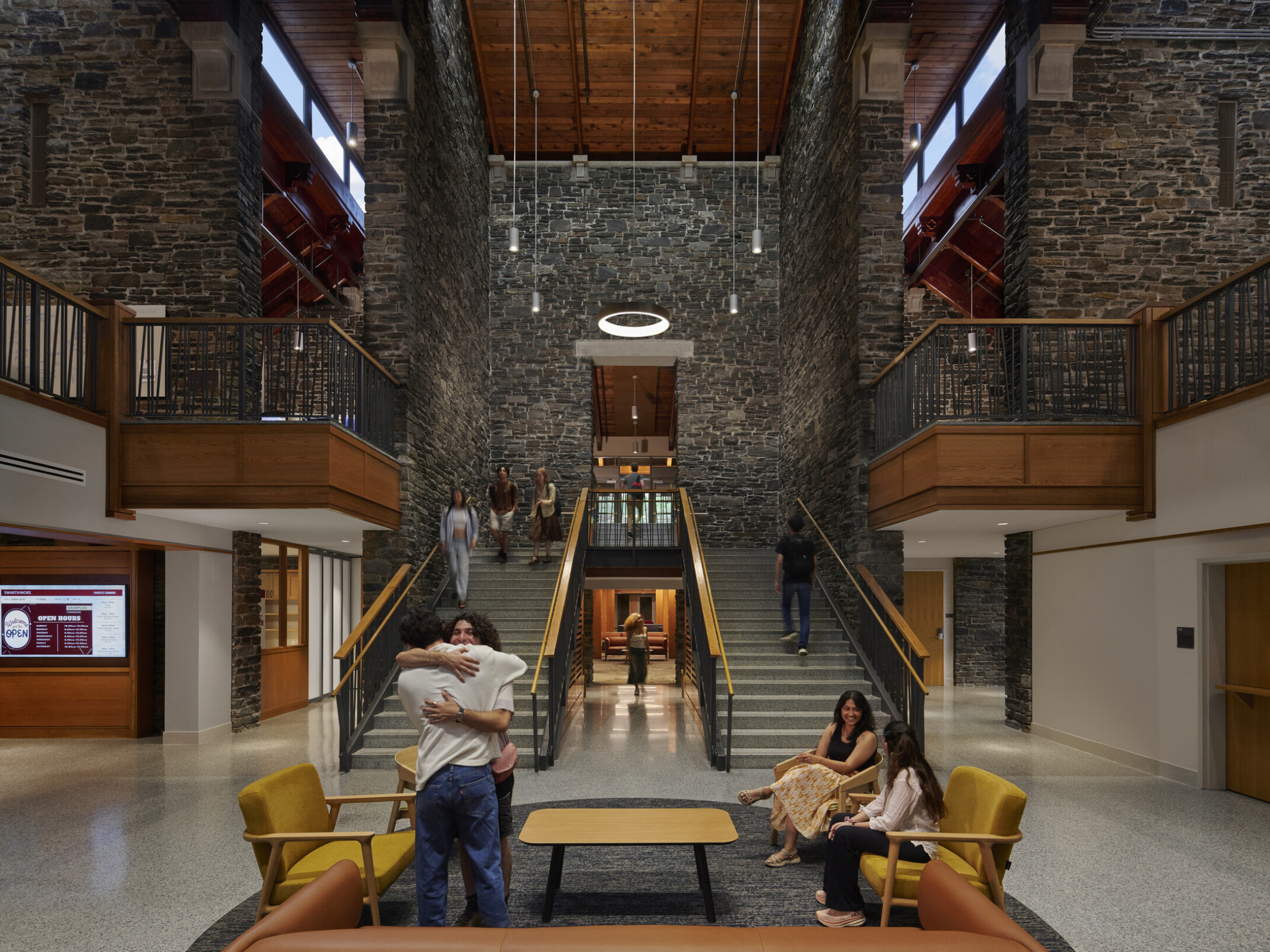 Broad staircase with wooden railings ascending to a second floor, flanked by stone walls, circular light fixture suspended from the ceiling. Seating area with yellow and brown chairs and a rectangular wooden coffee table on a speckled gray floor at the base of the stairs