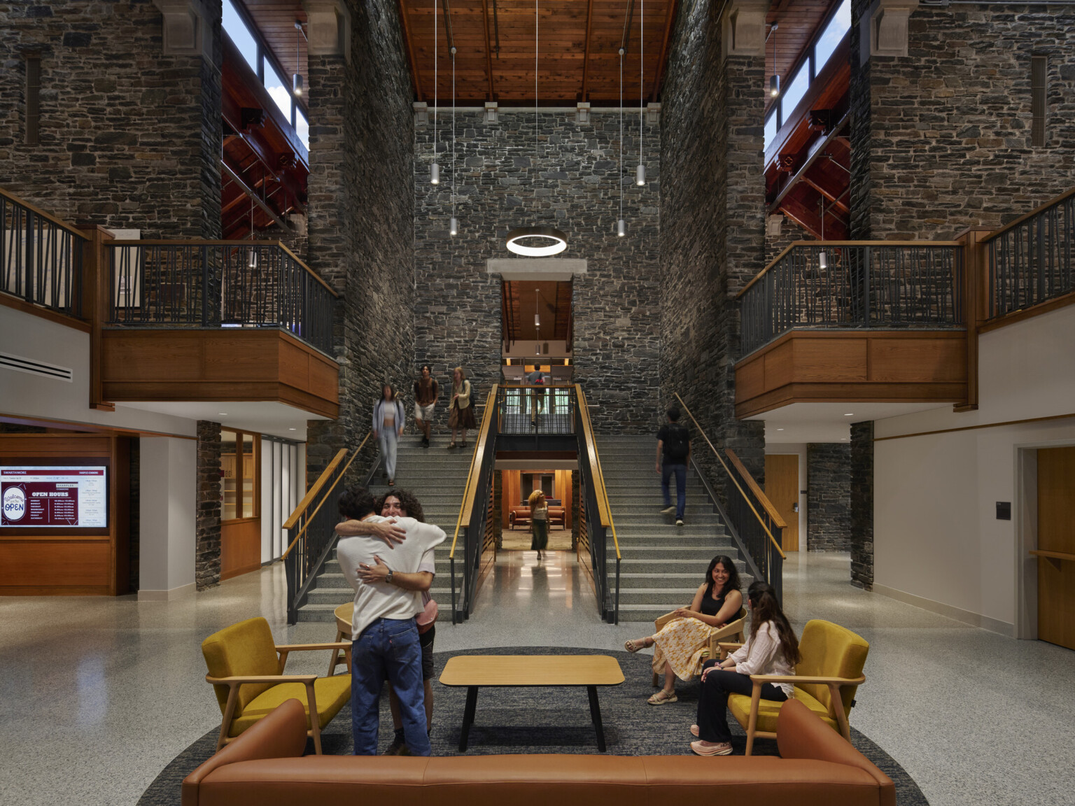 Broad staircase with wooden railings ascending to a second floor, flanked by stone walls, circular light fixture suspended from the ceiling. Seating area with yellow and brown chairs and a rectangular wooden coffee table on a speckled gray floor at the base of the stairs