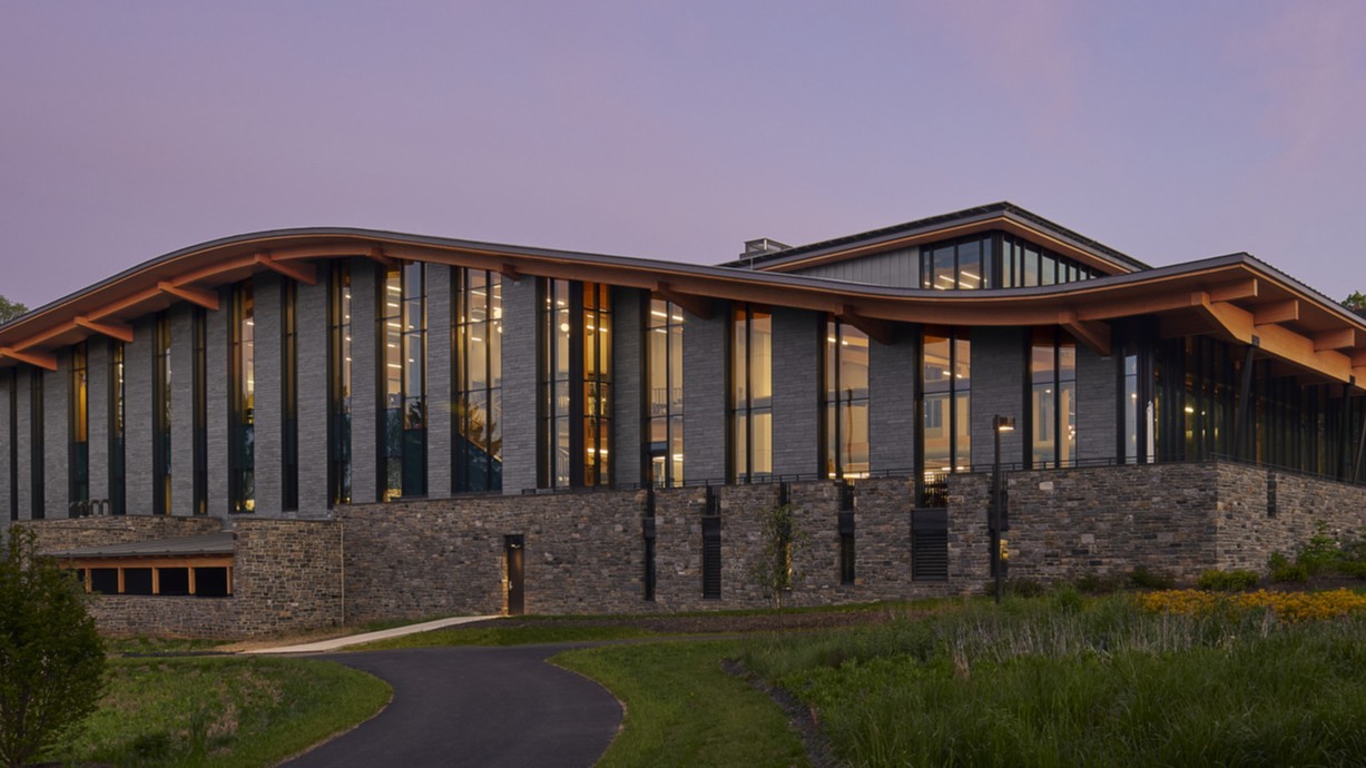 Stone and glass facade on university building with curving roof and overhang illuminated from within in the evening, mass timber architecture, what is mass timber construction