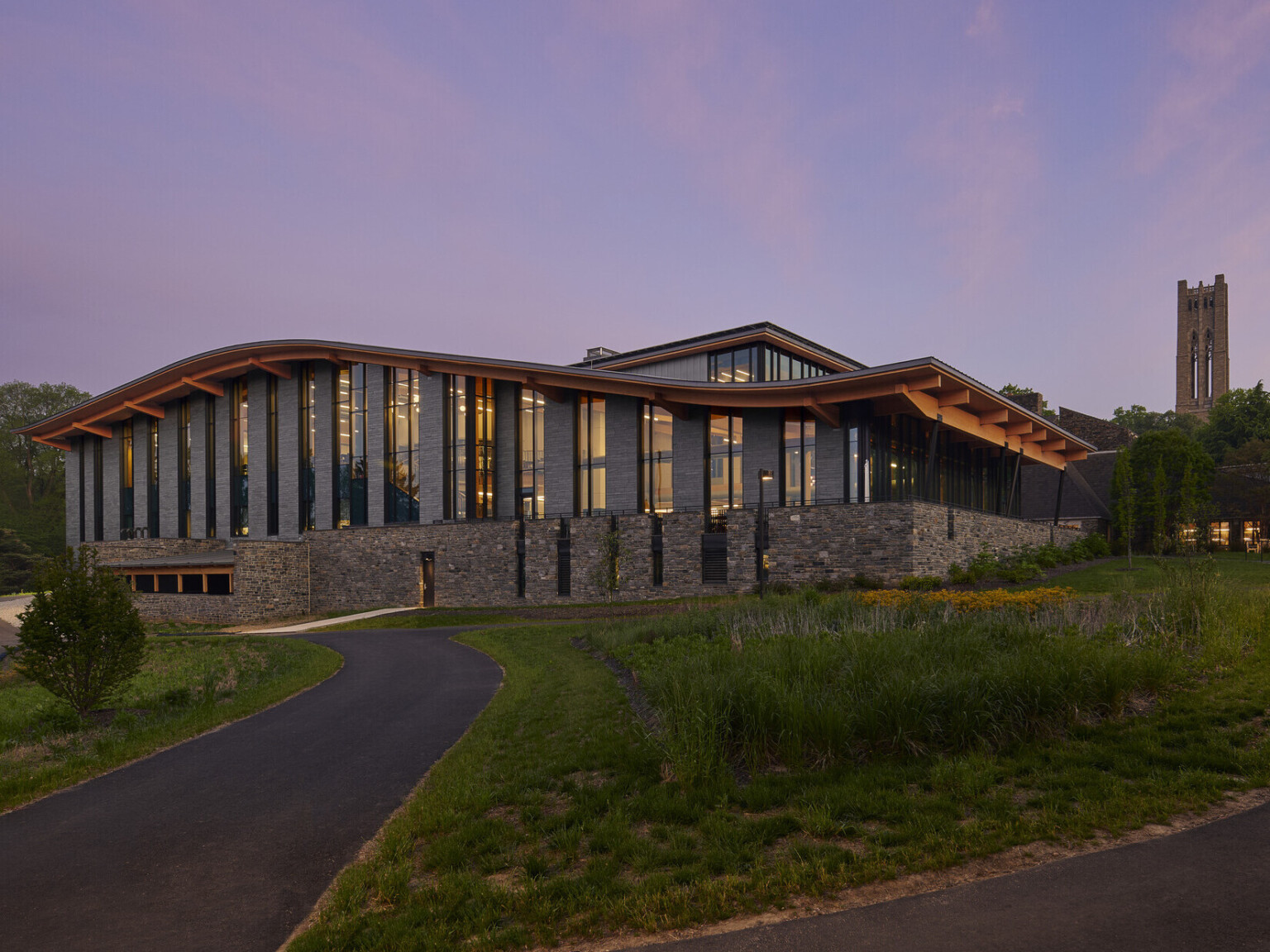 Stone and glass facade on university building with curving roof and overhang illuminated from within in the evening, mass timber architecture, what is mass timber construction