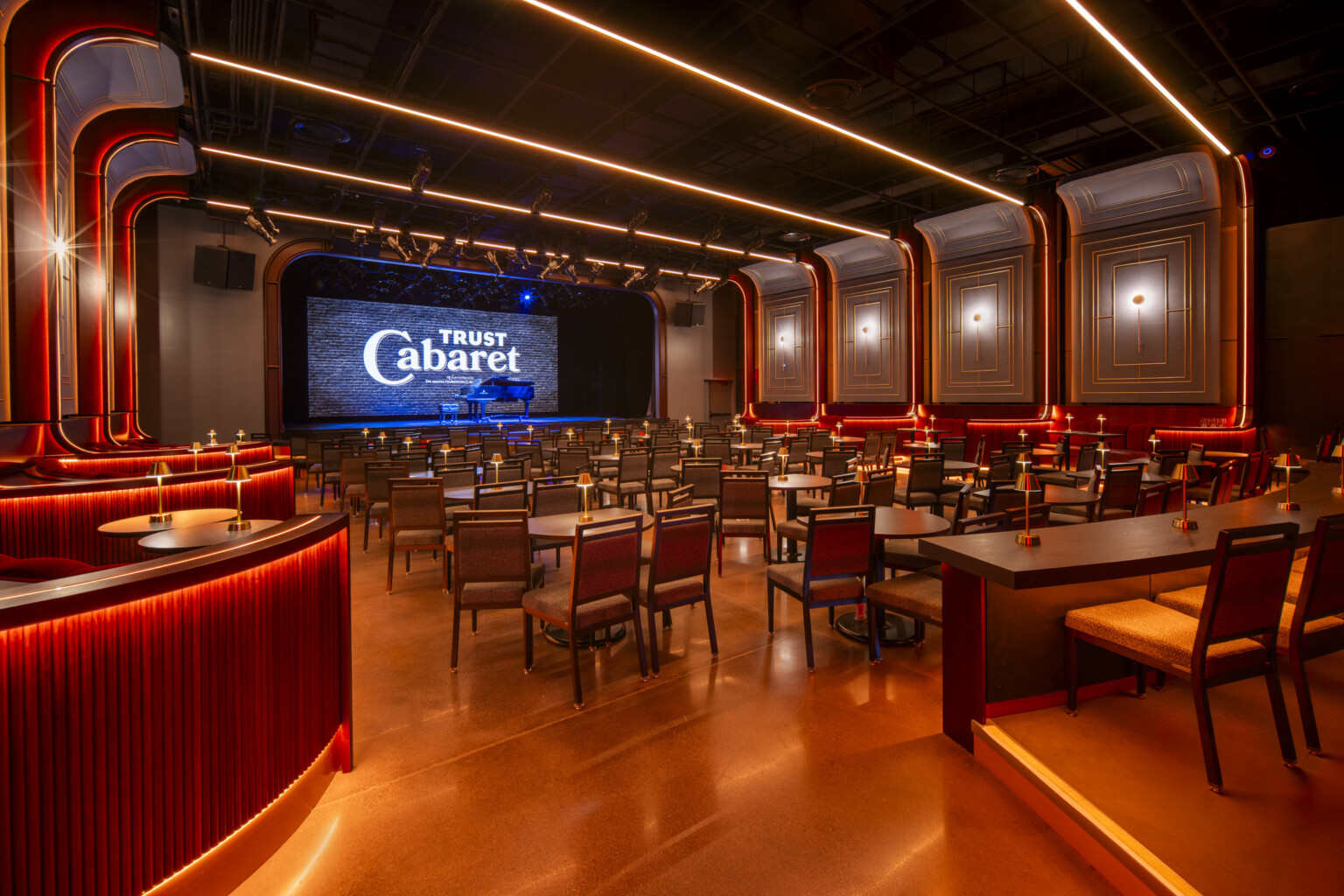 Lounge filled with tables and chairs facing a stage with a piano in the middle. Ceiling and walls accented by neon lights. Lounge filled with tables and chairs facing a stage with a piano in the middle. Ceiling and walls accented by neon lights.