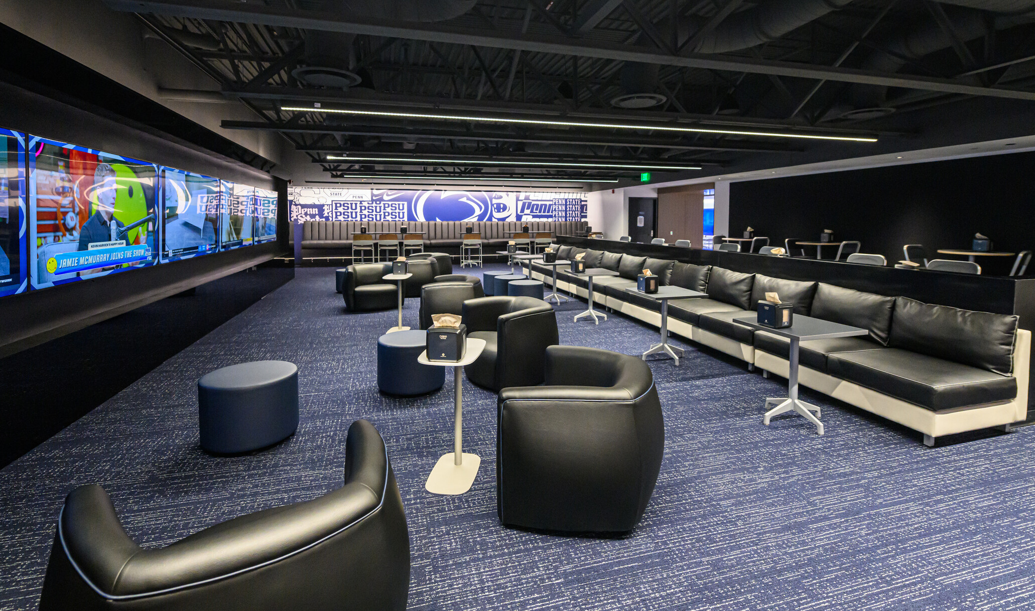 Penn State’s Greenberg Indoor Sports Complex lounge space; dimly lit room with blue carpets and black varied leather seating; large screens lining the walls of the space