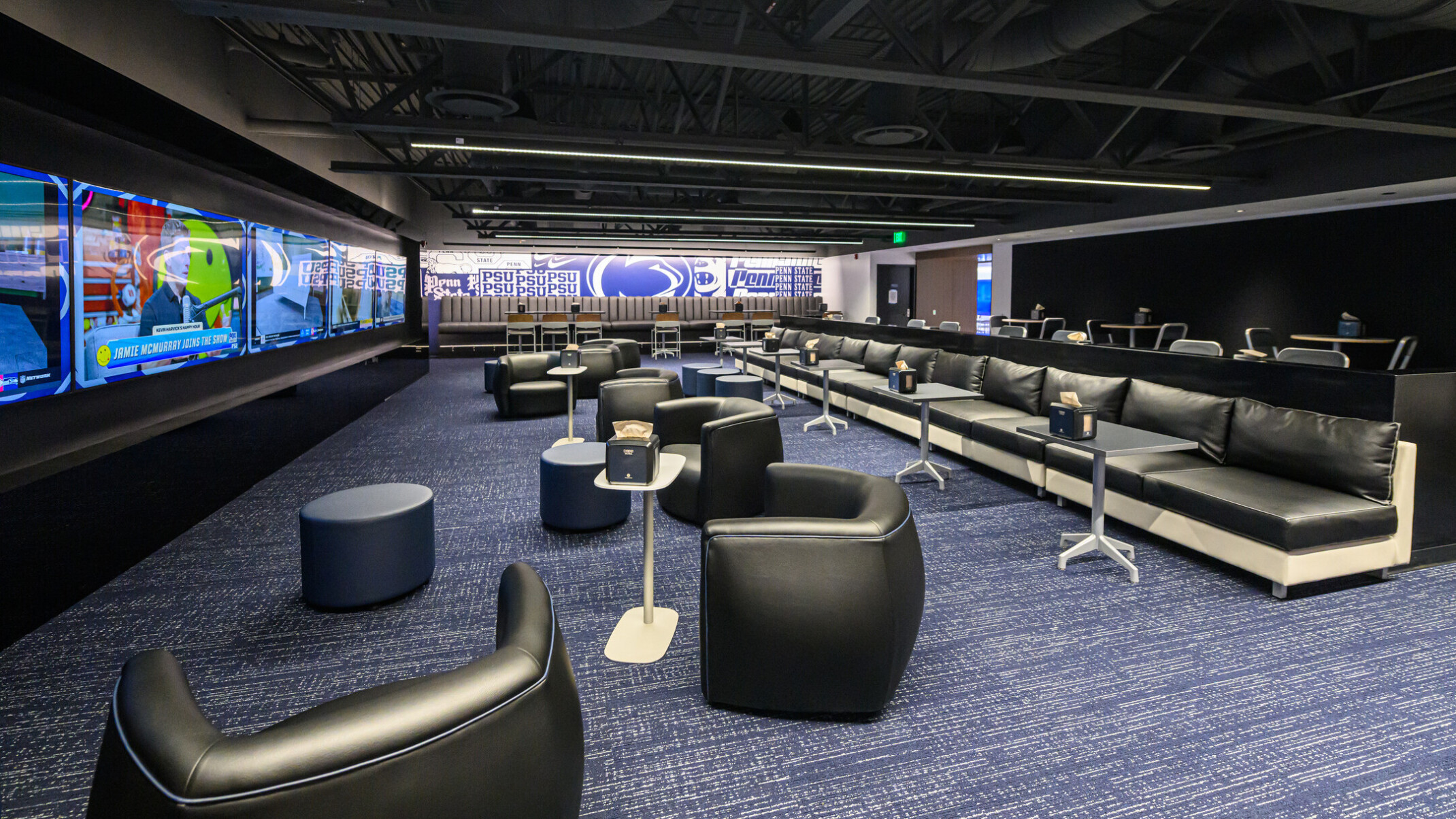 Penn State’s Greenberg Indoor Sports Complex lounge space; dimly lit room with blue carpets and black varied leather seating; large screens lining the walls of the space