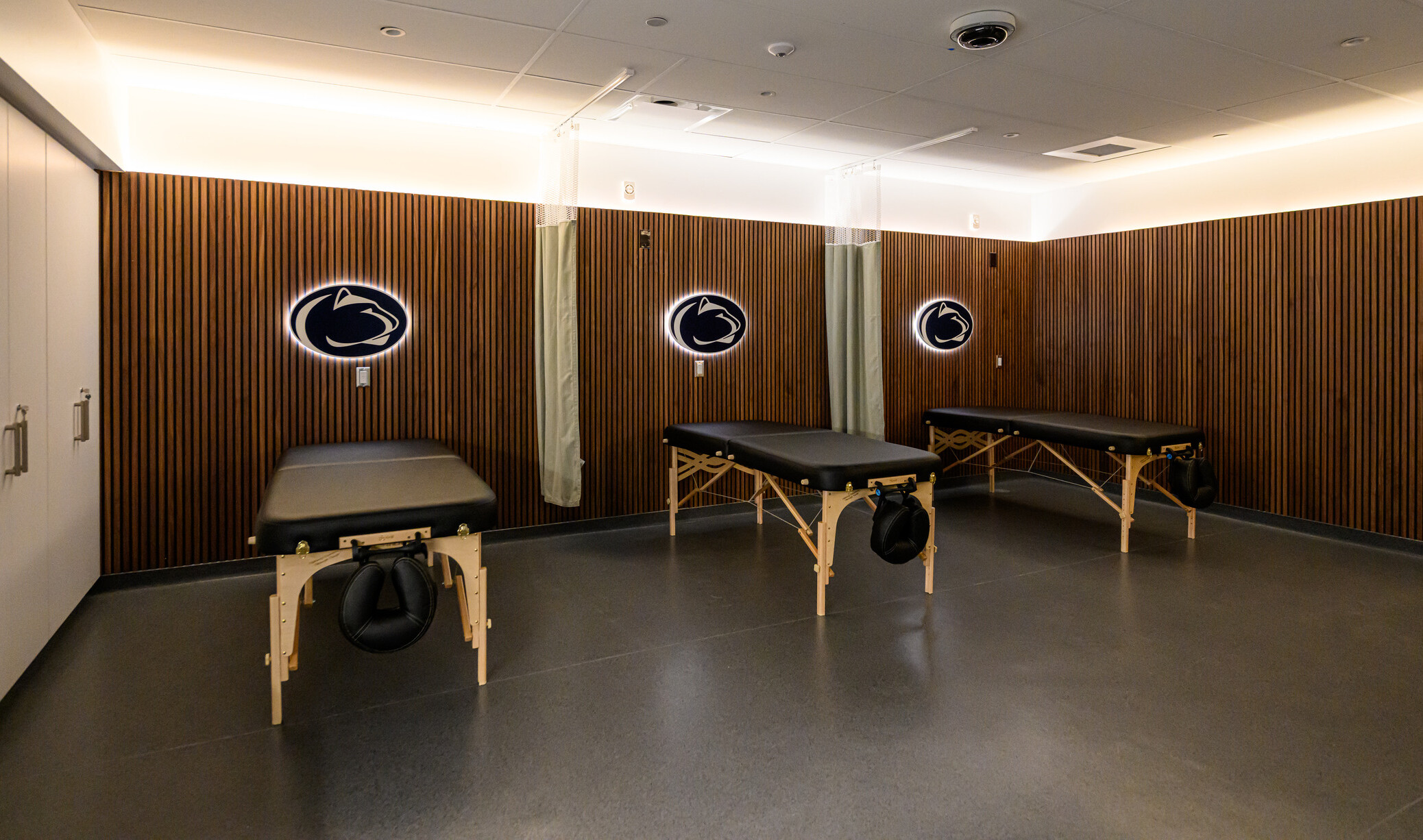 Penn State’s Greenberg Indoor Sports Complex treatment area; three black exam tables throughout the room with penn state logo above each
