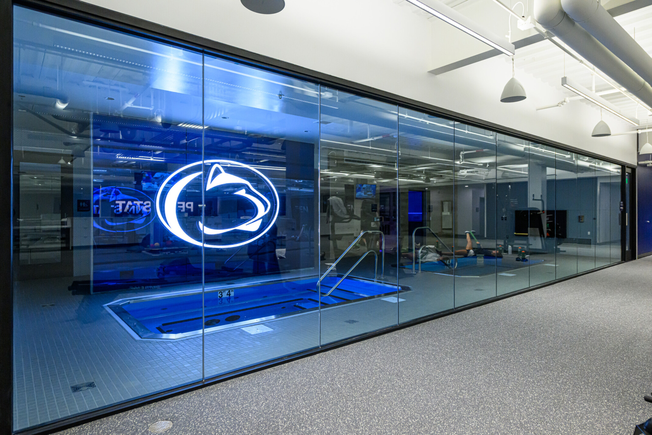 Penn State’s Greenberg Indoor Sports Complex recovery pool; large LED pen state logo above the small pool room; image pov outside the room looking in through glass walls