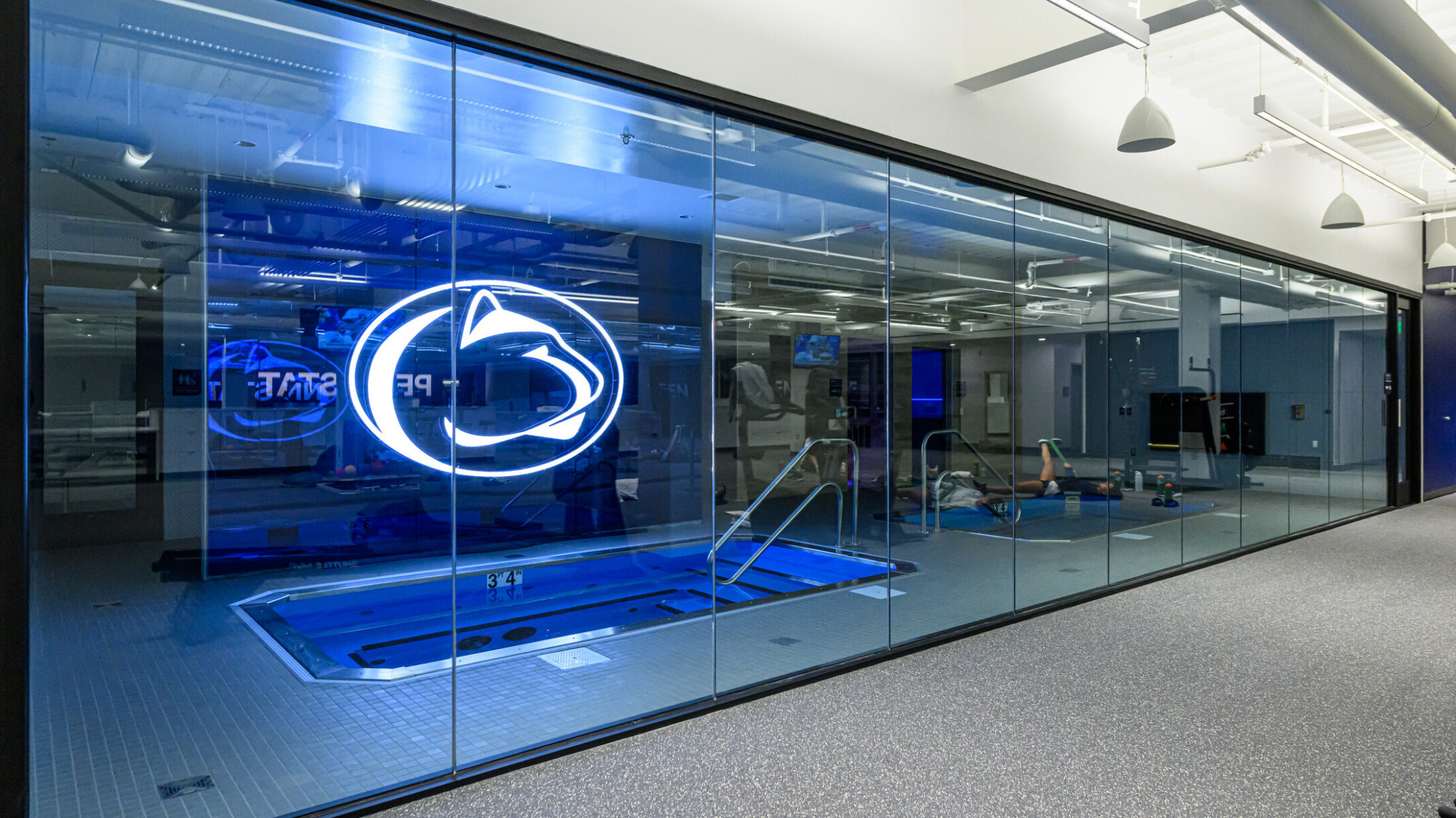 Penn State’s Greenberg Indoor Sports Complex recovery pool; large LED pen state logo above the small pool room; image pov outside the room looking in through glass walls