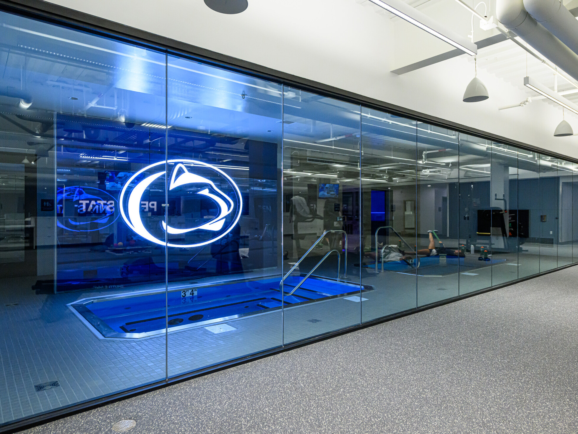 Penn State’s Greenberg Indoor Sports Complex recovery pool; large LED pen state logo above the small pool room; image pov outside the room looking in through glass walls