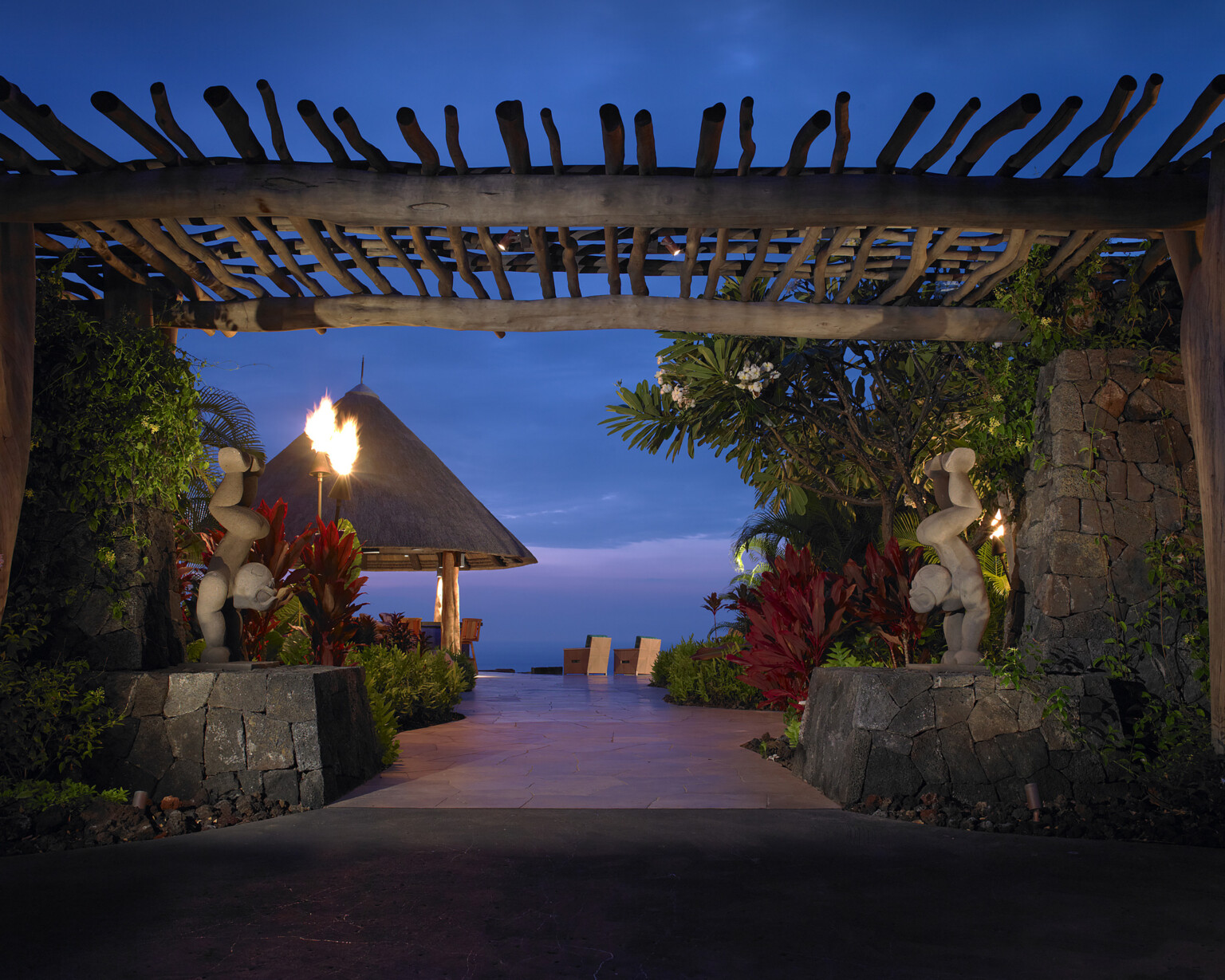 nighttime view of the ocean from a resort in Hawaii, natural wood canopy covers the walkway while sculptures, tiki torch, and traditional hut line the way