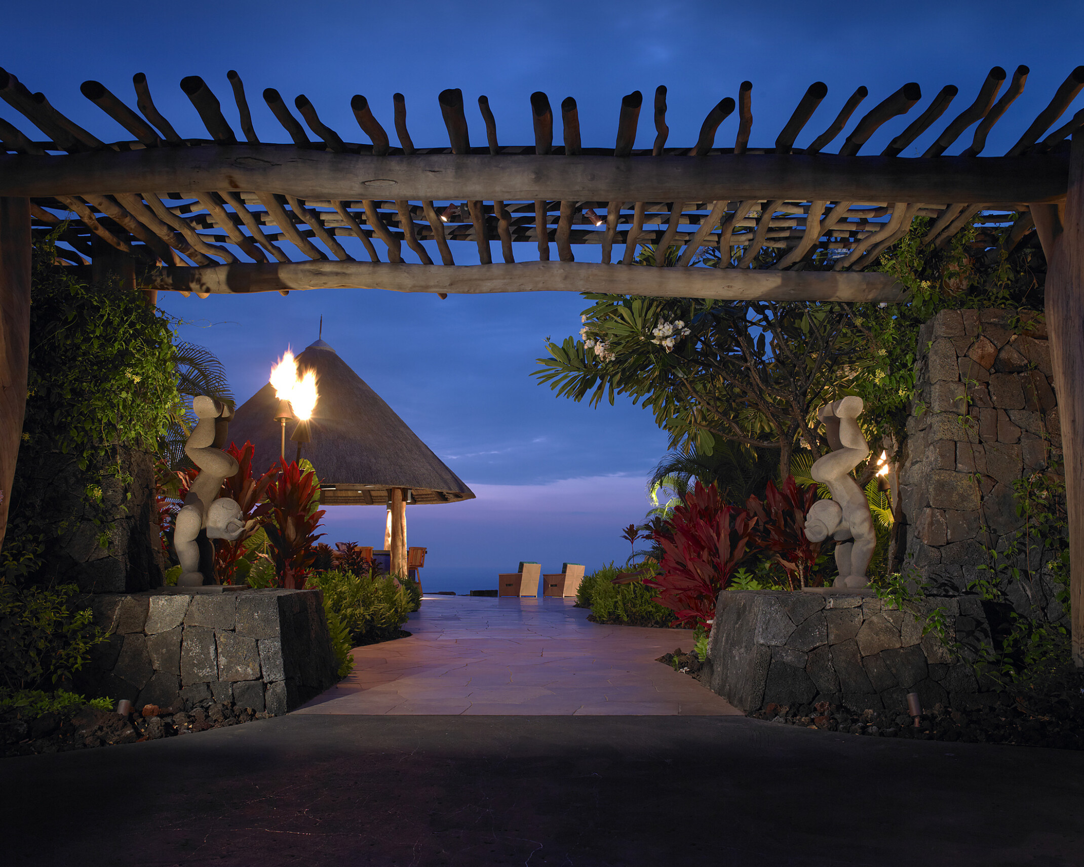 nighttime view of the ocean from a resort in Hawaii, natural wood canopy covers the walkway while sculptures, tiki torch, and traditional hut line the way