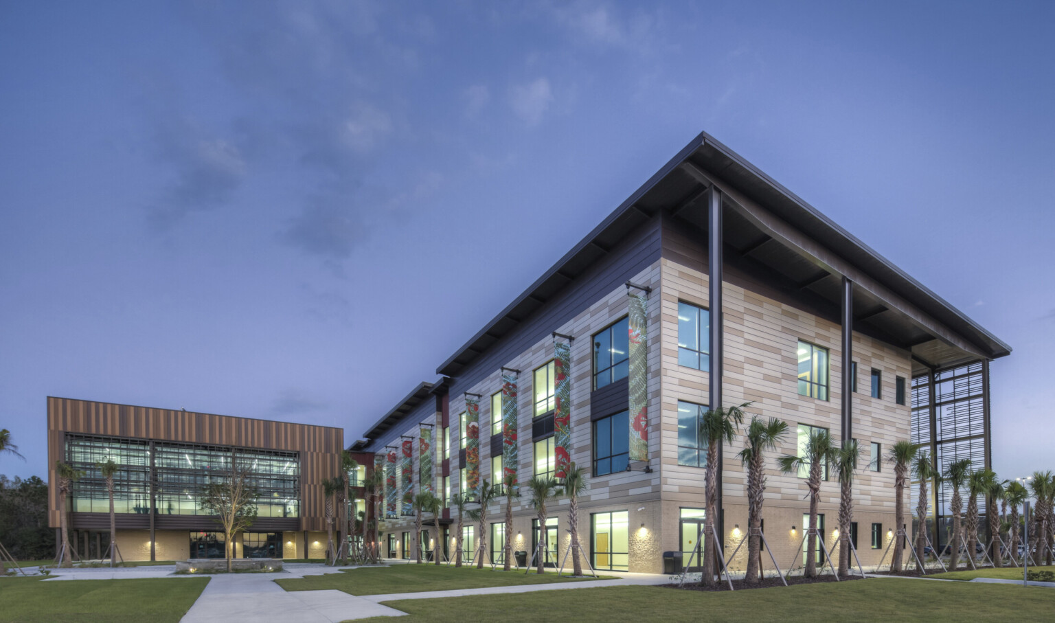 Exterior corner view of stone building lined with palm trees. Brown paneled building behind with double height windows