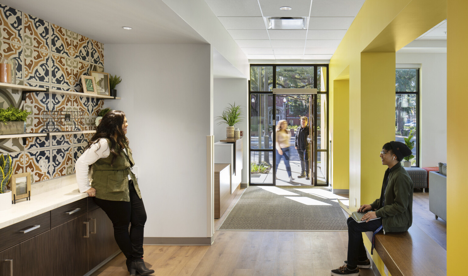 Hallway left of yellow wall to entry. White room towards the door behind recessed dark wood counter with muraled backsplash