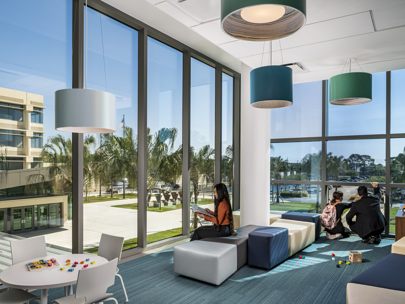 courthouse family area, high-ceilings, floor-to-ceiling windows fill all walls provide natural light, soothing blues and greens for flooring, modular seating areas, barrel lamps overhead with blue and green shades