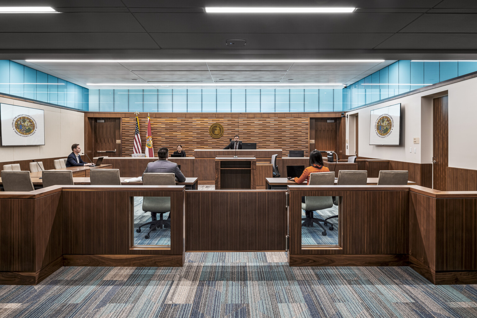 four people seated in family courtroom, turquoise-tinted clerestory windows run across three walls below the ceiling providing natural light, darker wood panels on back wall, podium, desks, dividers and gates, geometric patterned low-pile carpet in soothing cool hues