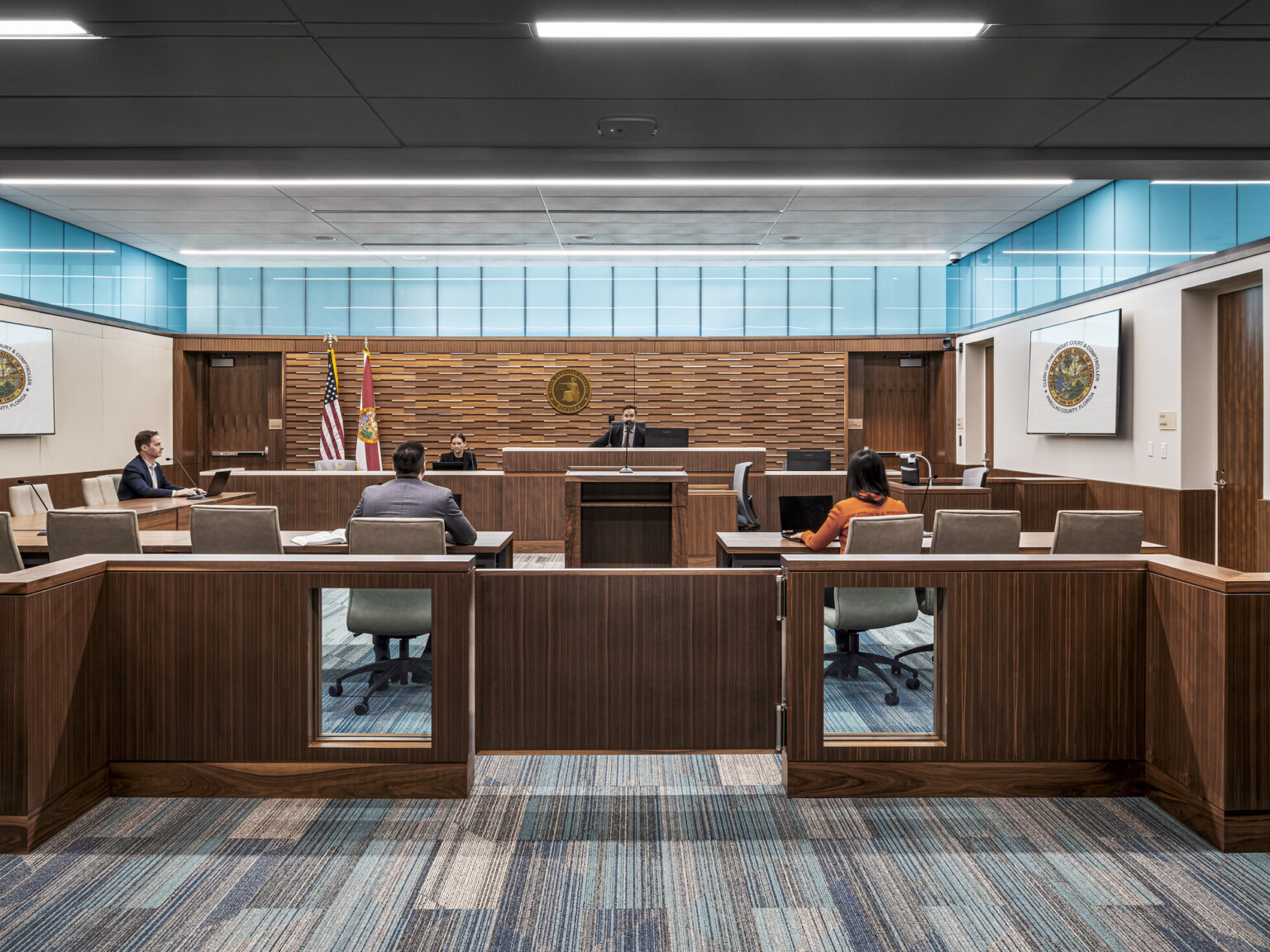 four people seated in family courtroom, turquoise-tinted clerestory windows run across three walls below the ceiling providing natural light, darker wood panels on back wall, podium, desks, dividers and gates, geometric patterned low-pile carpet in soothing cool hues
