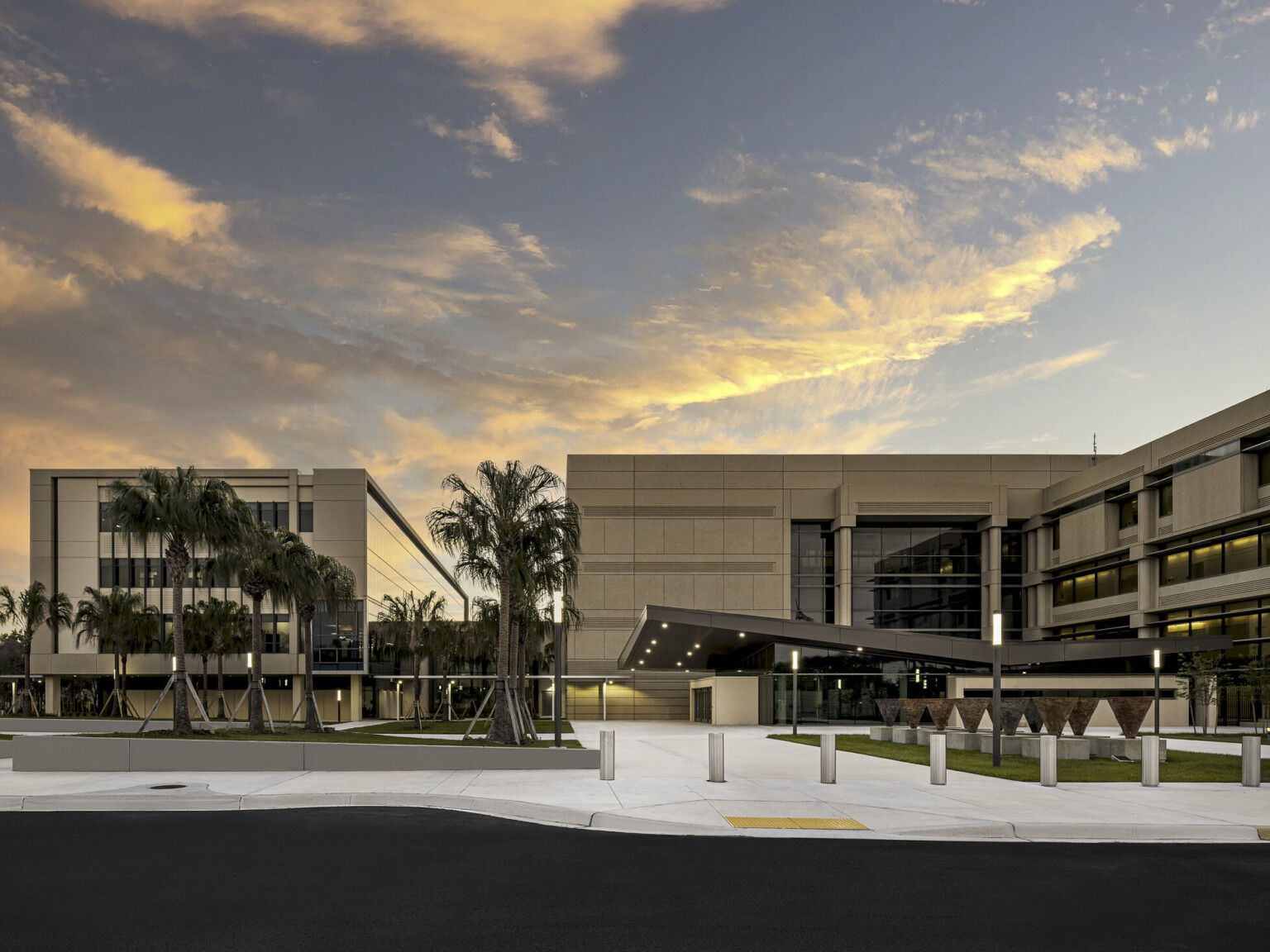 exterior view of building, beige concrete facade, modern steel supports, lighted walkway, palm tree, orange and pink clouds at dusk