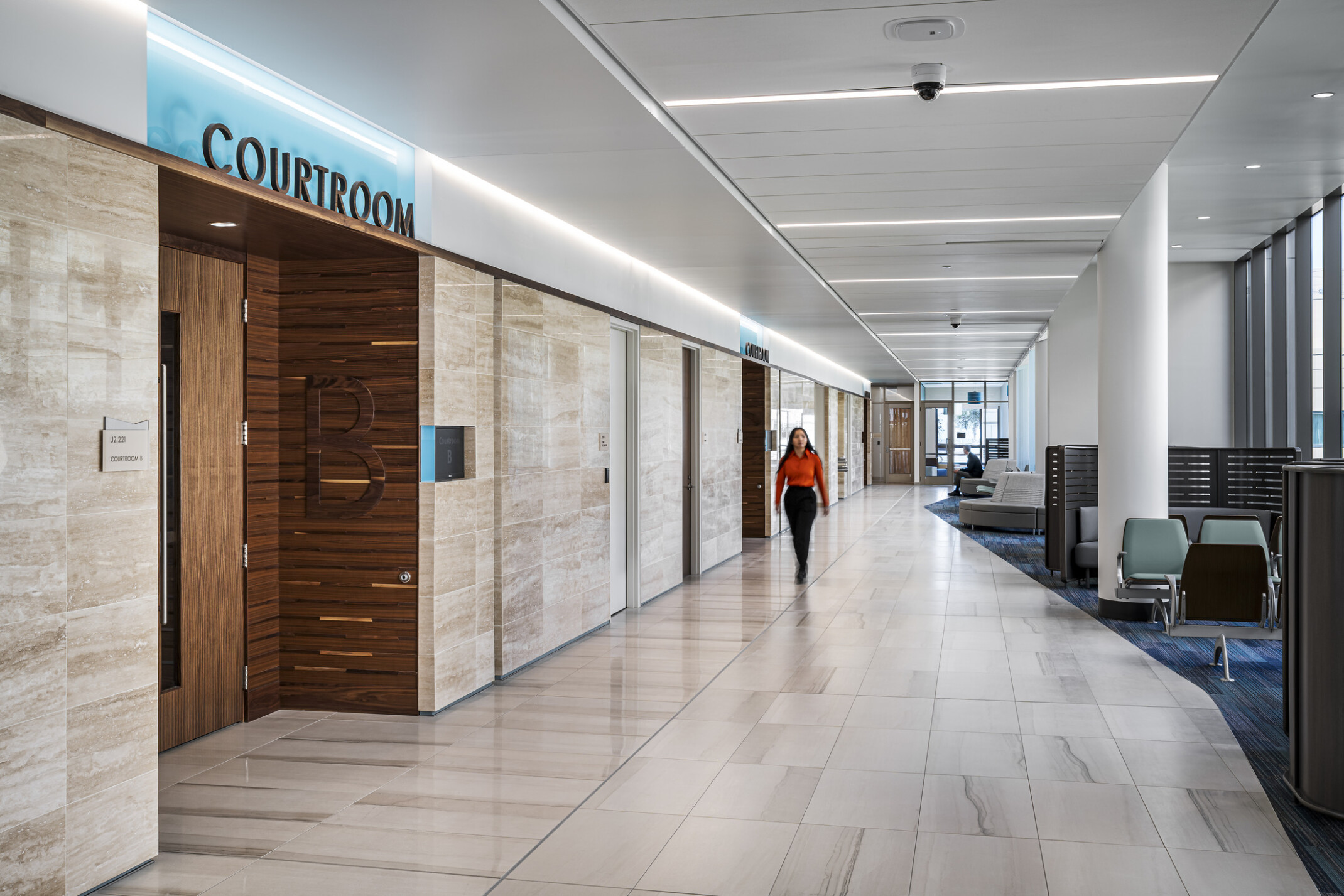 Woman dressed in a red long sleeve shirt walking down a hallway exterior to Courtroom B in the The Pinellas County Family Courts Annex; public seating and lounge space near windows.