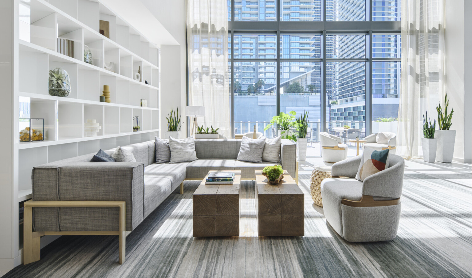 Bespoke white divider shelf in large group with gray, modern seating, wood block coffee table, and planters in double height glass room