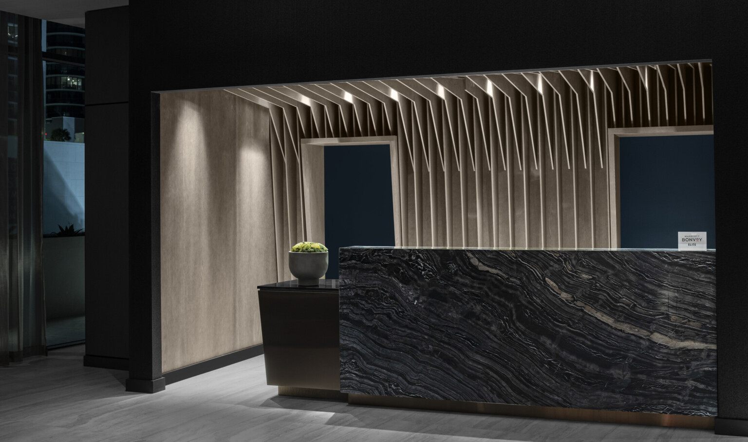 The AC Hotel reception desk, a dark stone slab with warm wood slats like palm fronds vertically emerge overhead in recessed space