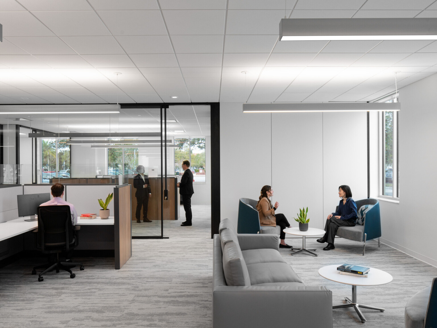 Office space waiting area filled with grey couches and blue chairs to the side of a desk space with desks separated by low partitions