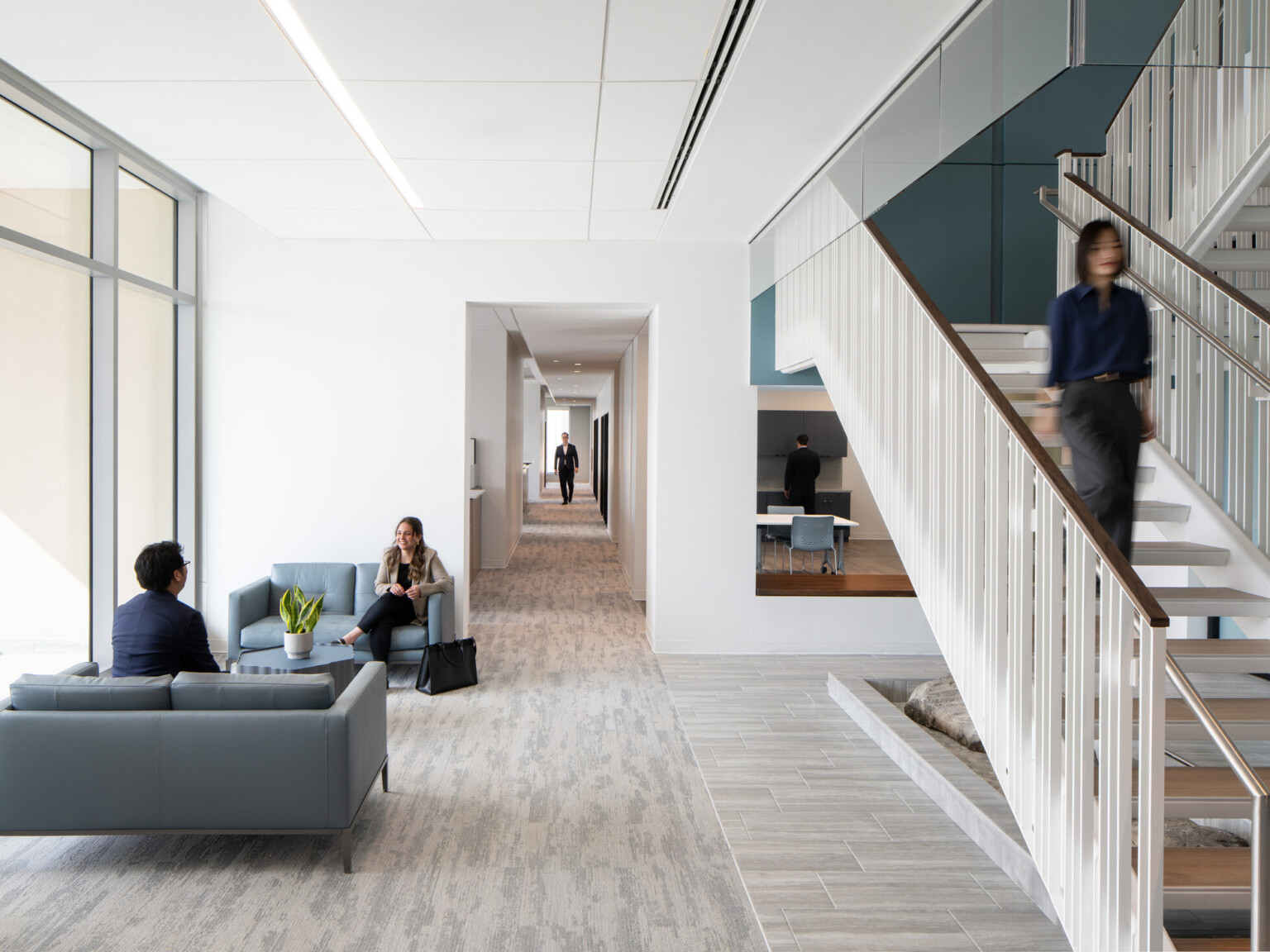 Office interior looking down a hallway with a modern white staircase to the right, grey flooring, and blue furniture in a waiting area
