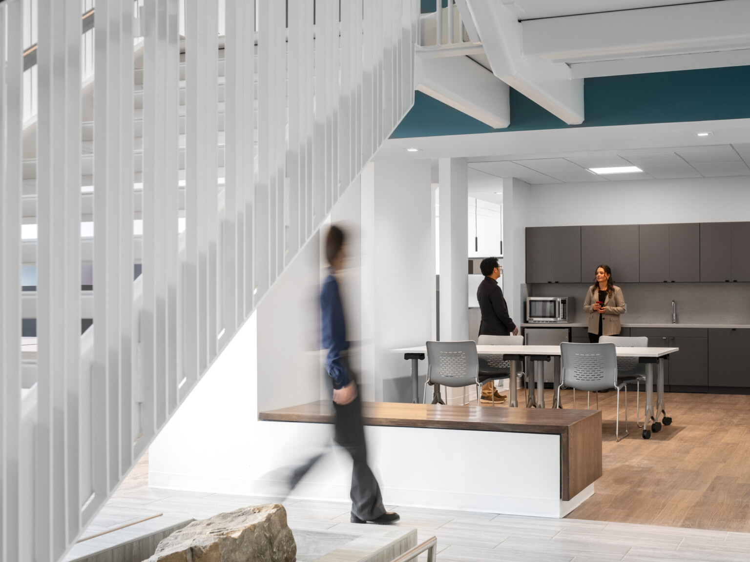 Office breakroom with dark grey cabinets, white tabletop with grey chairs, and blue accents with a staircase to the left