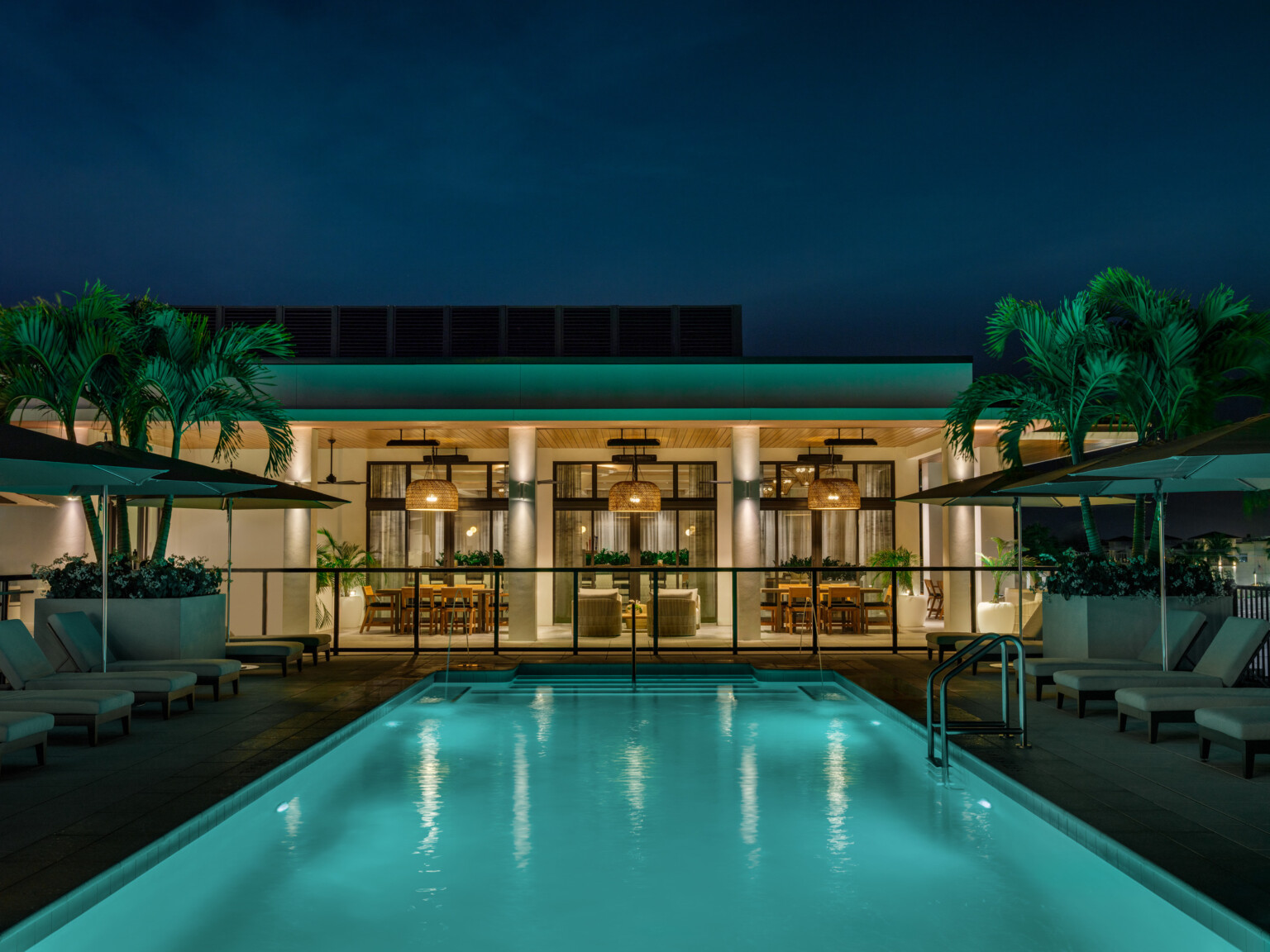 Nighttime rendering of a hotel pool showing turquoise waters in front of an outdoor patio with round light fixtures handing from the ceiling