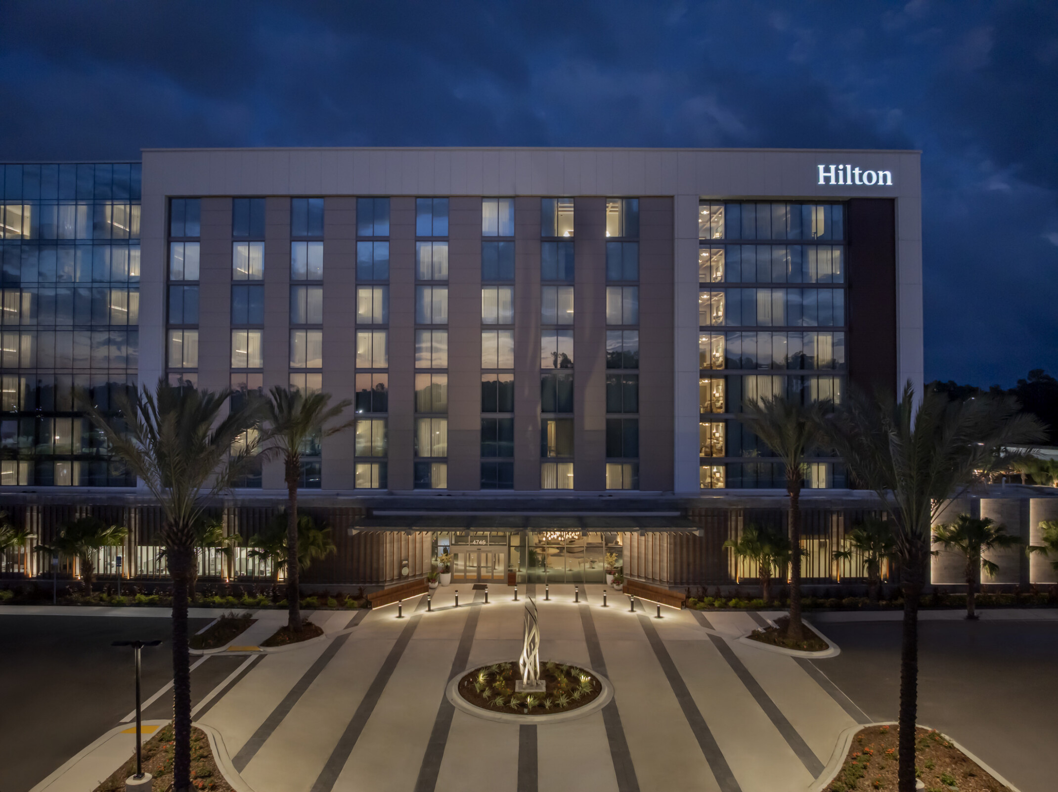 Straight-on view of Hilton Jacksonville main entry with glowing porte cochere, roundabout sculpture, and branded Hilton signage anchoring the façade