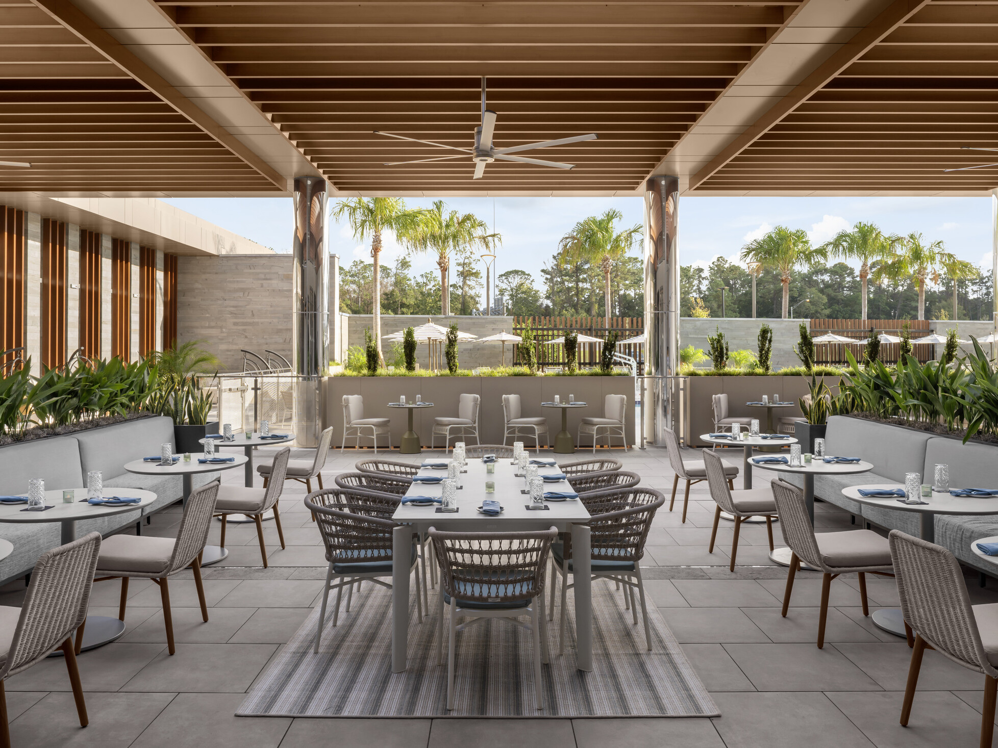Outdoor restaurant seating at Hilton Jacksonville featuring banquet and family-style tables beneath a wood slat ceiling, with light gray furnishings accented in soft blue
