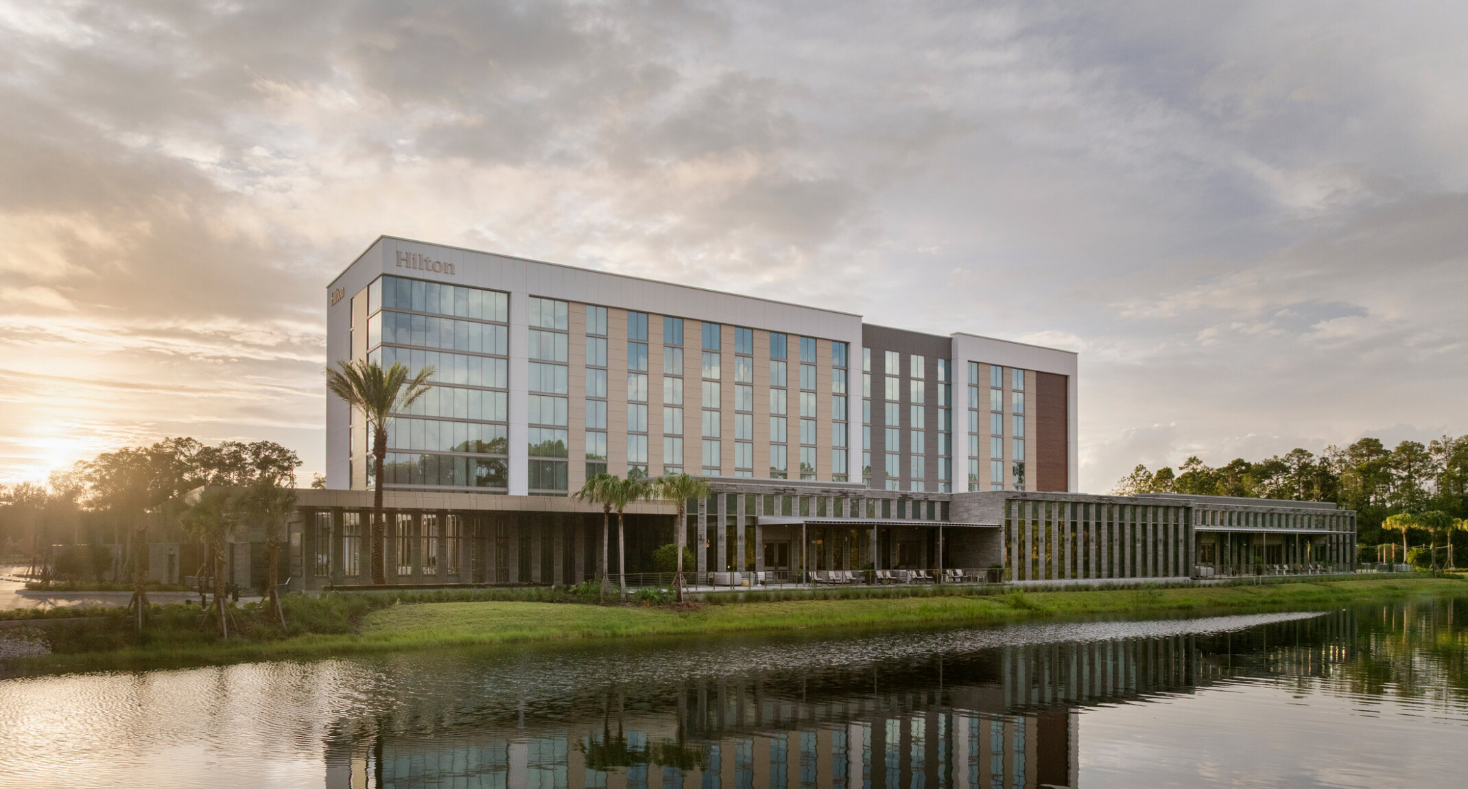 Hilton Jacksonville at Mayo Clinic façade overlooking the main pond, featuring expansive glass curtain wall, covered outdoor seating beneath a deep overhang, and pedestrian access to the conference center entrance.