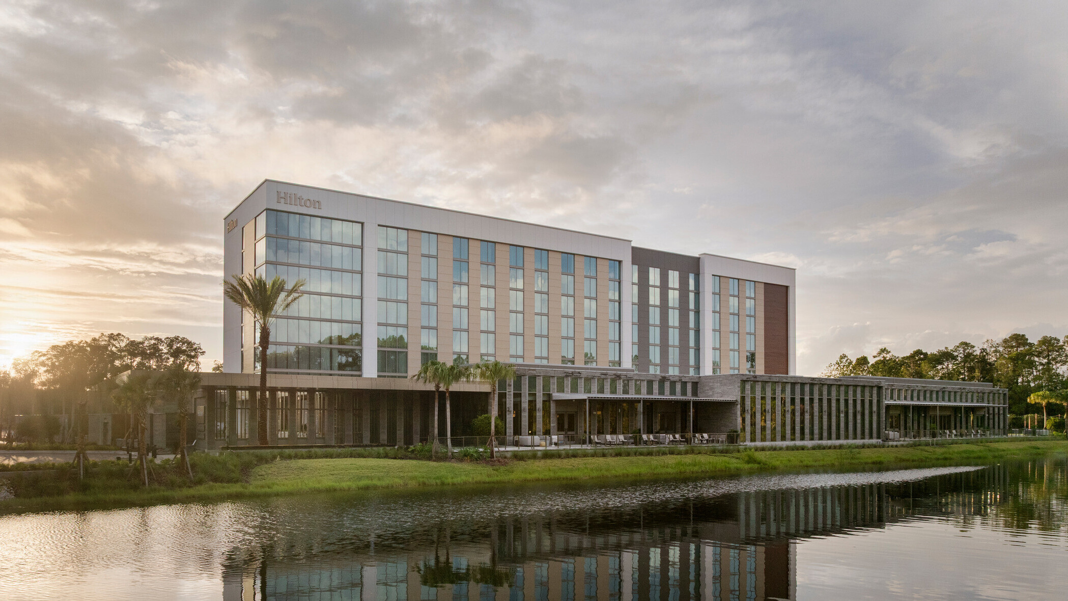 Hilton Jacksonville at Mayo Clinic façade overlooking the main pond, featuring expansive glass curtain wall, covered outdoor seating beneath a deep overhang, and pedestrian access to the conference center entrance.